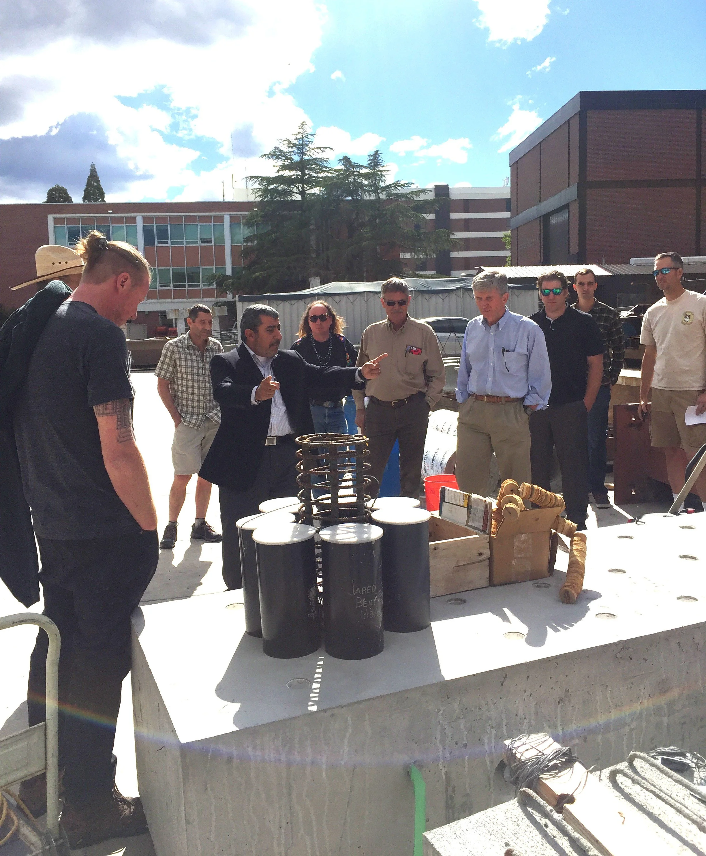 A group of people gathered outdoors around a table where a man is demonstrating objects, possibly at an educational event, with several onlookers listening.