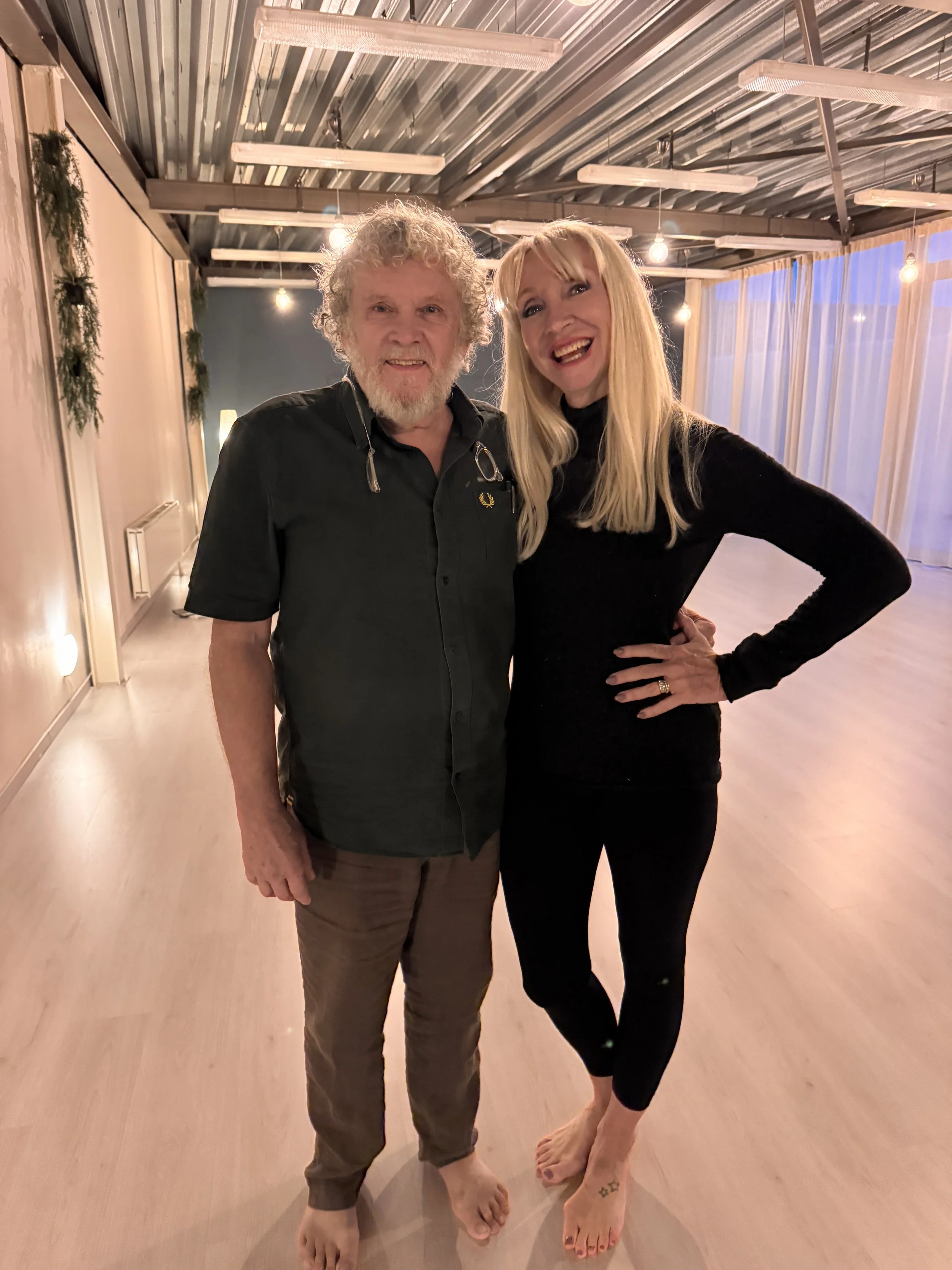 Due persone sorridenti, un uomo anziano con capelli ricci e barba bianca, una donna con capelli lunghi biondi e gonne nere, che intentano a una festa in un ambiente ben illuminato con luci soffuse.