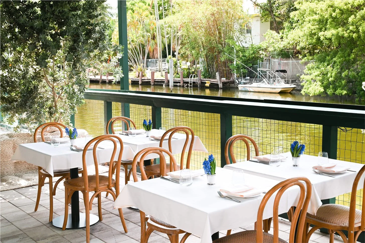 Outdoor restaurant patio overlooking a waterway with trees, a boat, and docks. Four tables with white tablecloths, blue flowers in vases, glasses, napkins, and silverware, set for dining.