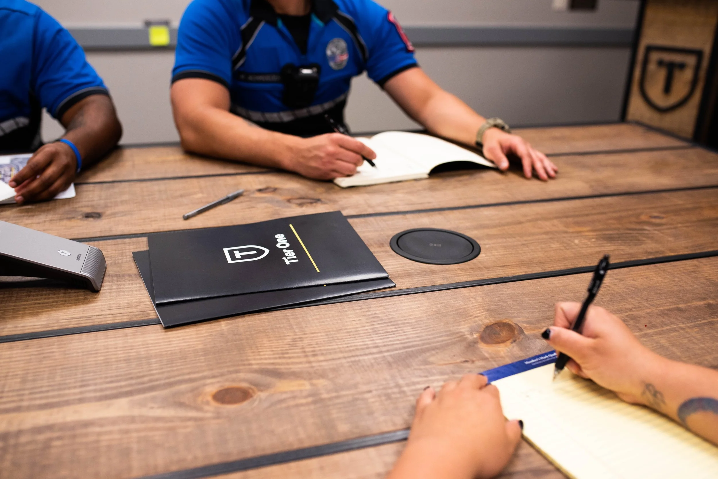 People sitting around a wooden conference table, taking notes and having a meeting, with folders and a wireless speaker on the table.