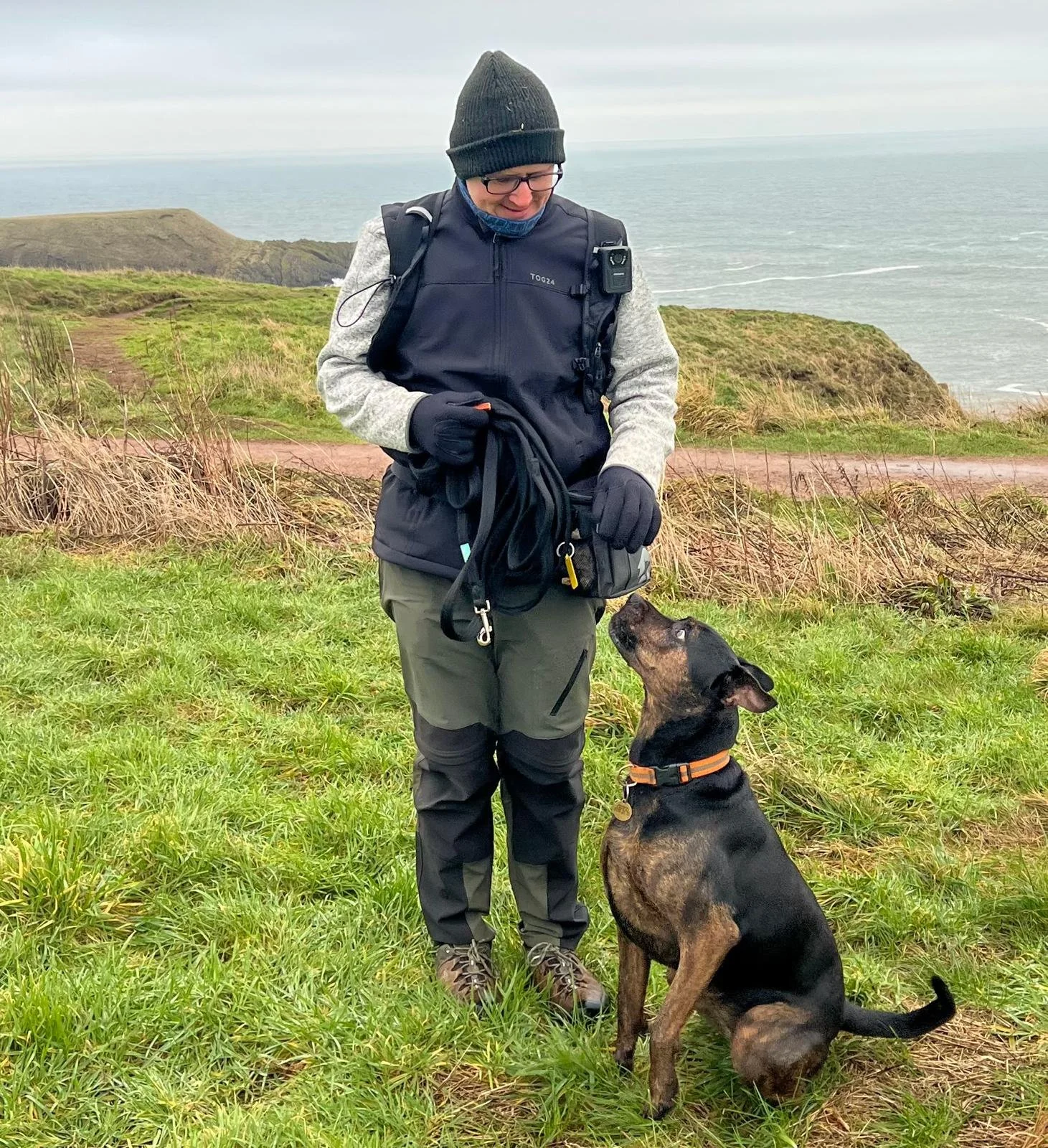 A person wearing outdoor gear standing on grass with a dog sitting in front, looking up at them, overlooking a coastal landscape with grassy hills and the ocean in the background.
