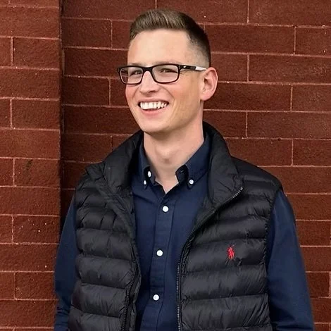A young man with glasses smiling and standing against a red brick wall, wearing a dark blue shirt and a black puffy vest.