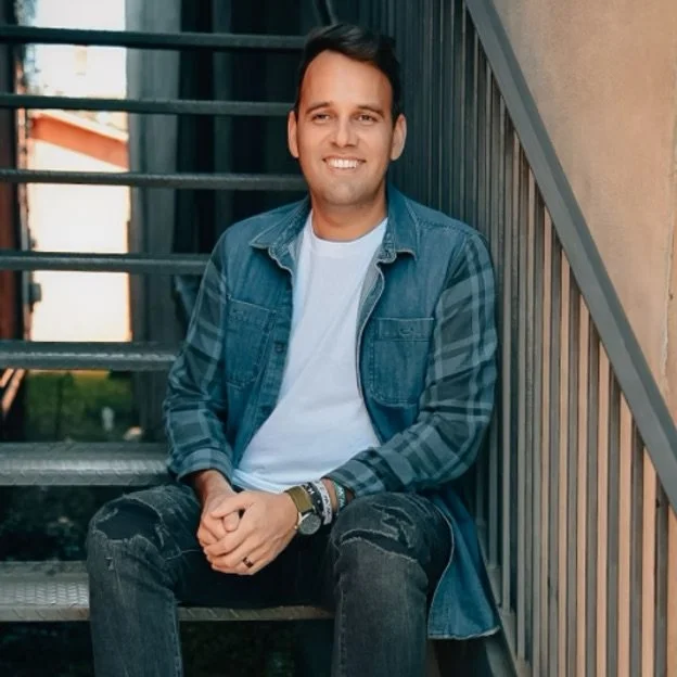 A young man sitting on outdoor stairs, smiling at the camera, wearing a denim jacket over a white t-shirt.