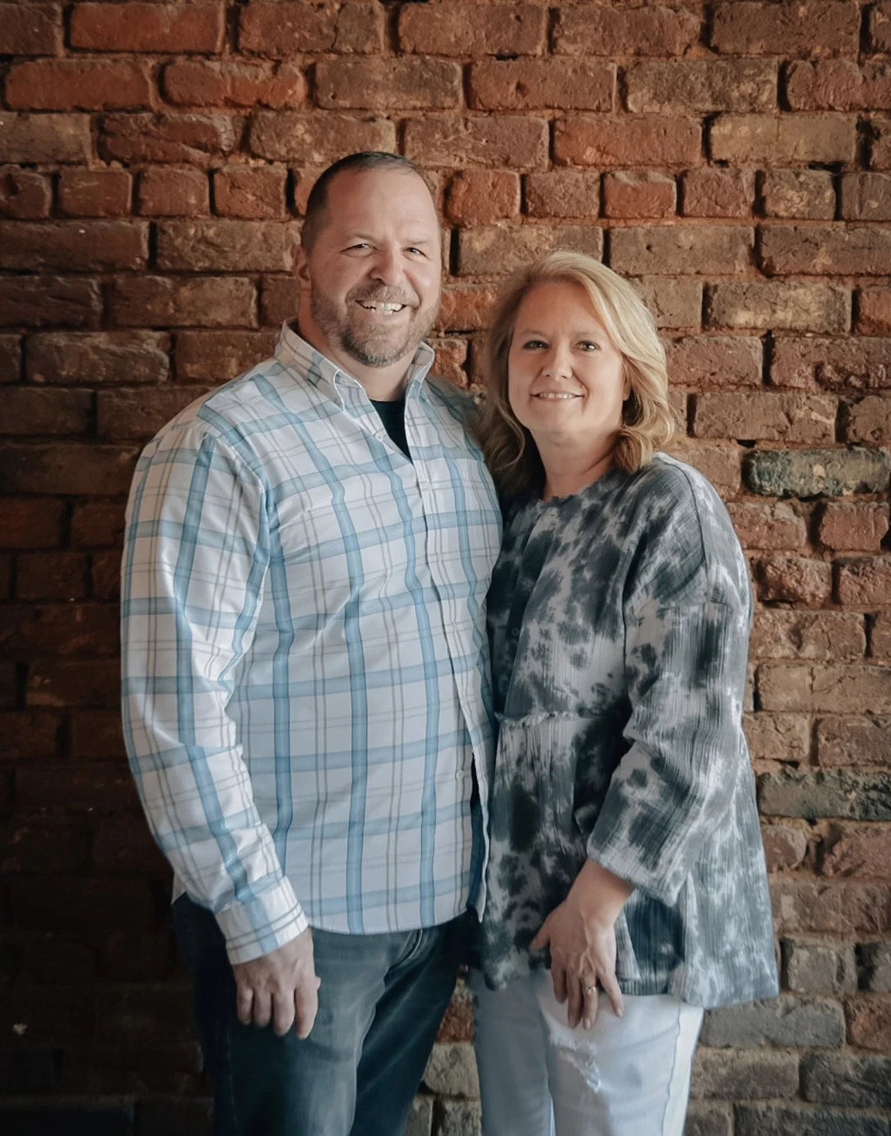 A smiling man and woman standing close together against a red brick wall, with the man wearing a blue plaid shirt and jeans, and the woman wearing a black and white tie-dye patterned blouse and white pants.