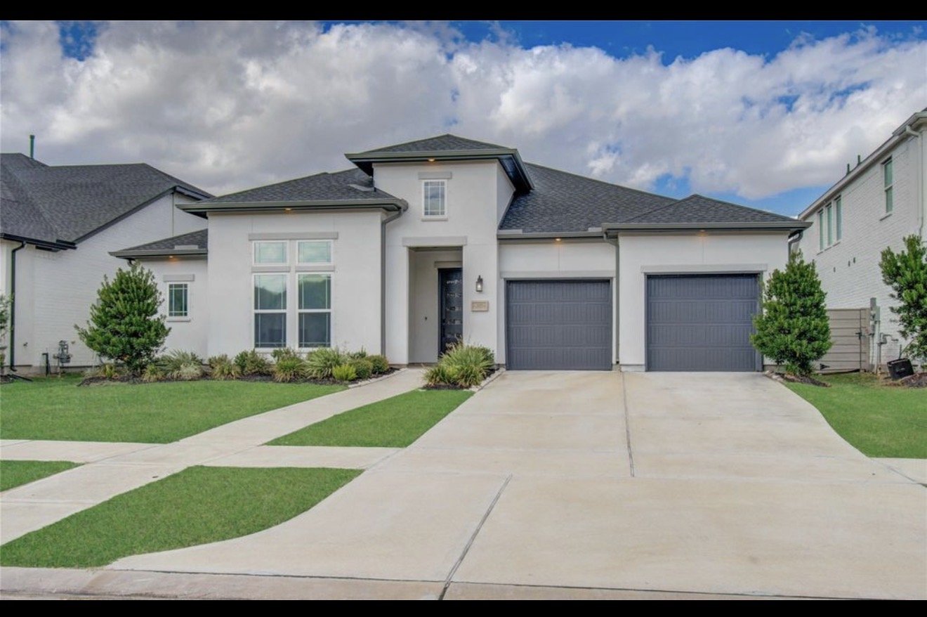 Front view of a modern two-story white house with dark gray garage doors, a black front door, well-kept lawn, small trees, and sidewalks.
