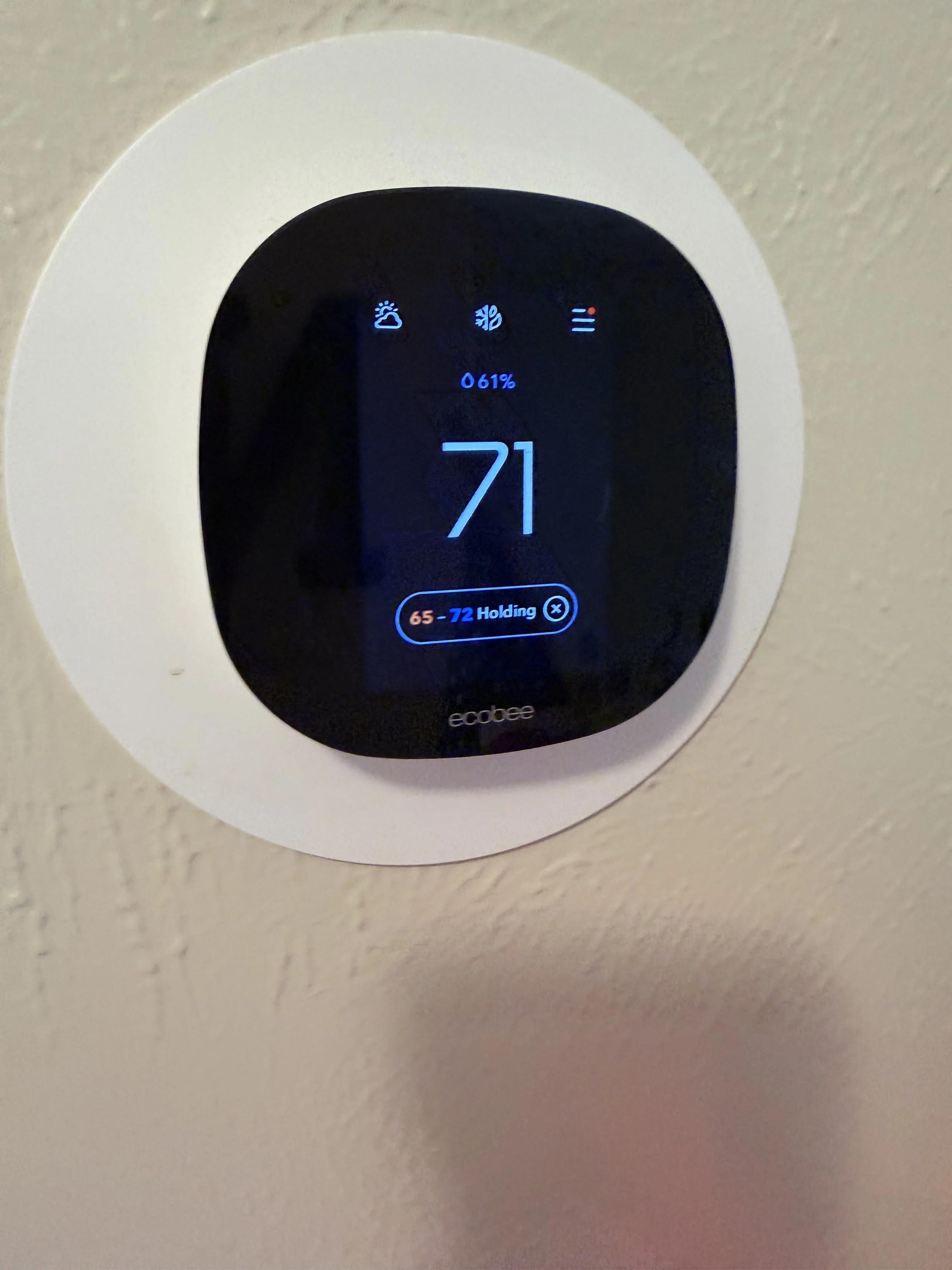 Digital thermostat on a wall displaying a temperature of 71 degrees Fahrenheit, with icons and settings for weather, humidity, and fan speed.