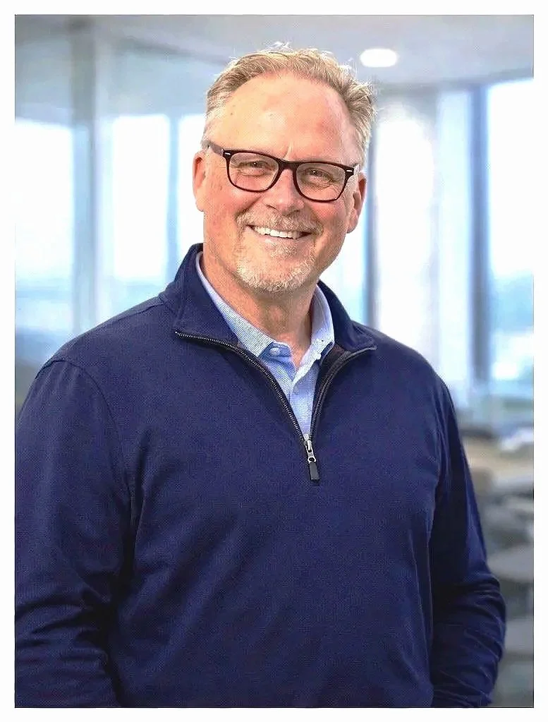 A smiling man wearing glasses, a blue quarter-zip pullover, and a light blue collared shirt, standing indoors with large windows in the background.