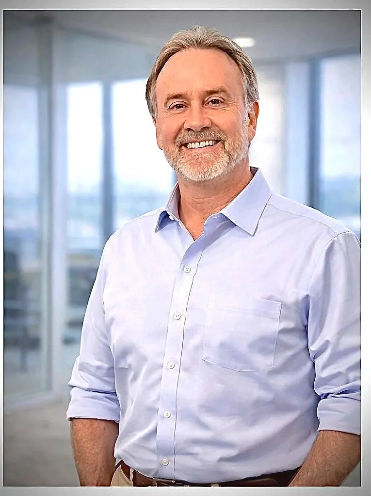 A middle-aged man with gray hair and a beard wearing a light blue button-down shirt, smiling and standing in a modern office with large windows.