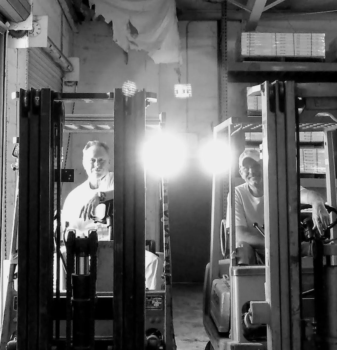 Two people operating forklifts in a warehouse, with shelves and boxes in the background, and bright lights behind them.