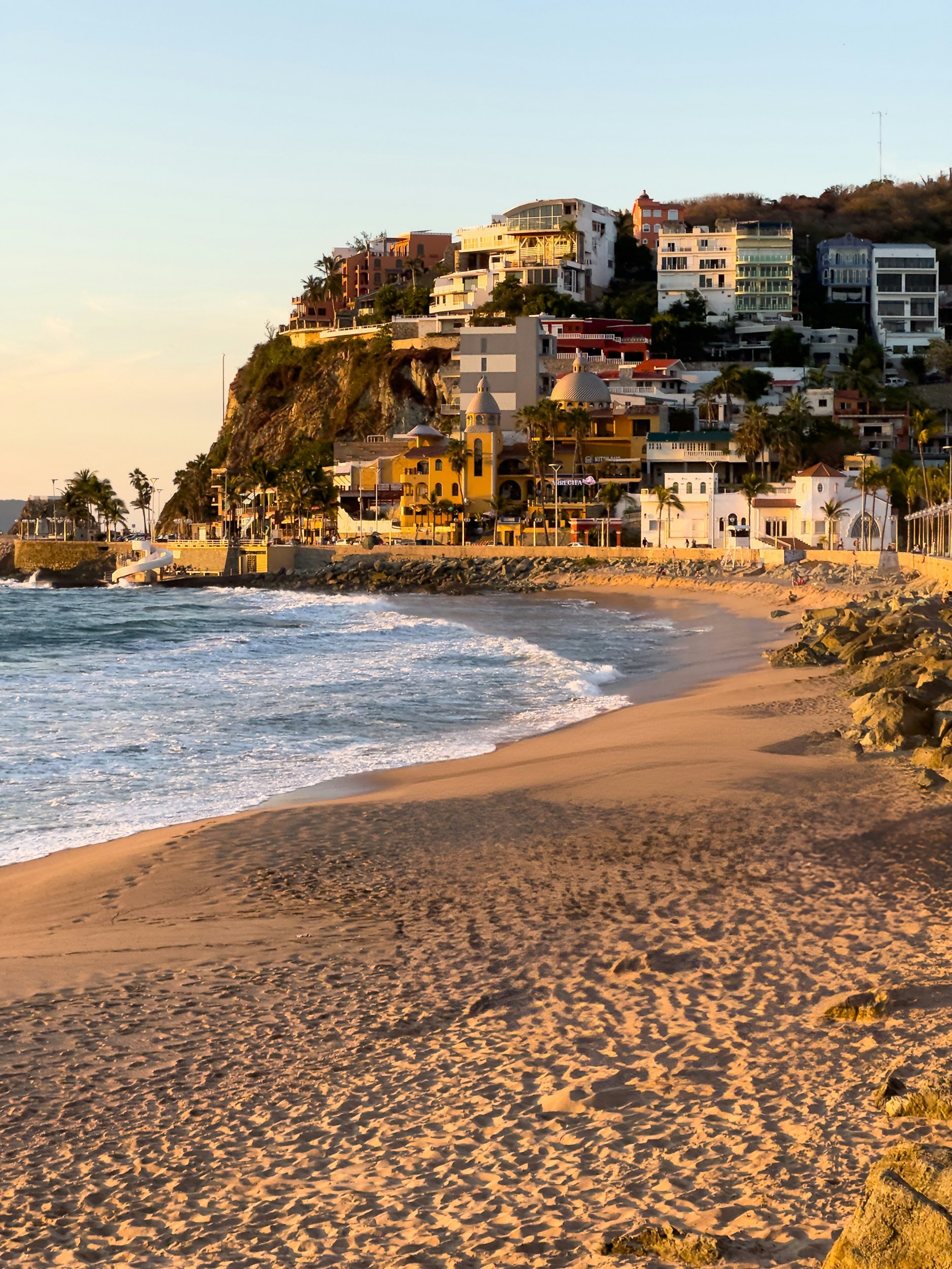 Sunset over a beach with buildings and houses on a hillside in the background.