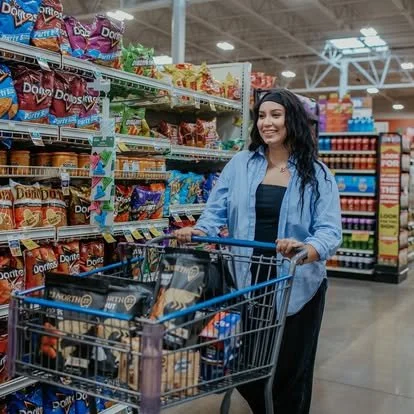 A woman shopping in a grocery store, pushing a cart with various snack bags including Doritos and other chips, standing in an aisle with shelves stocked with snack products.