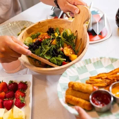Hands serving a mixed greens salad from a wooden bowl at a table with various other dishes.