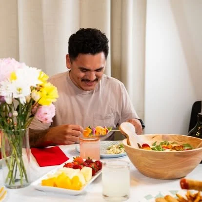 A man sitting at a table with a large vegetable and fruit salad, a glass of water, and a bowl of chips, with a vase of flowers nearby, smiling while holding a fork.