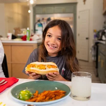 A young girl with long brown hair smiling and holding a sandwich inside a kitchen.