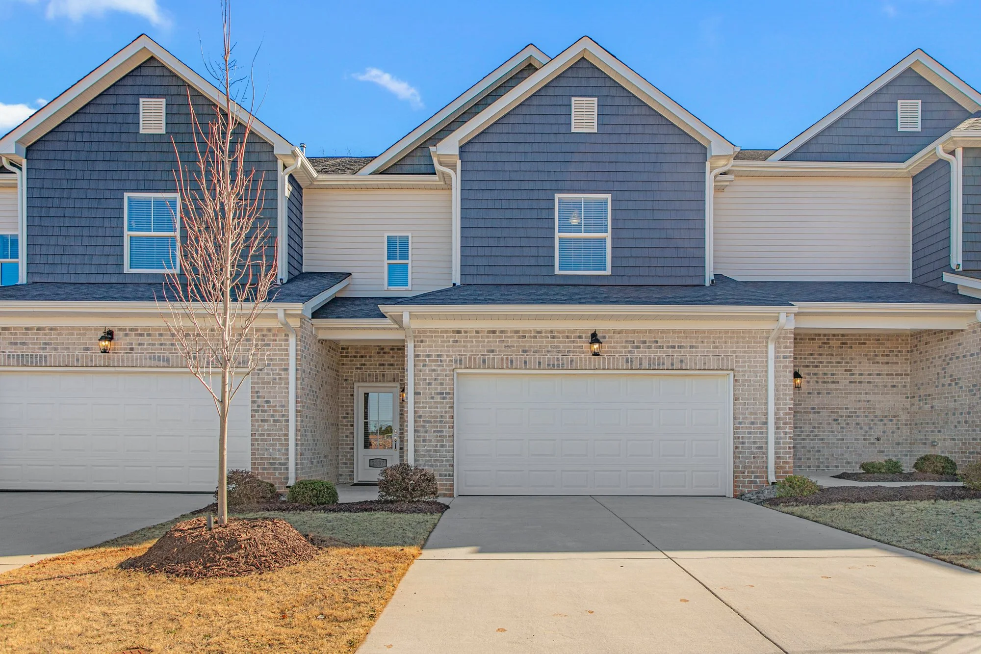 Modern townhouse with brick and siding exterior, two-car garage, leafless tree in front yard, and clear blue sky.