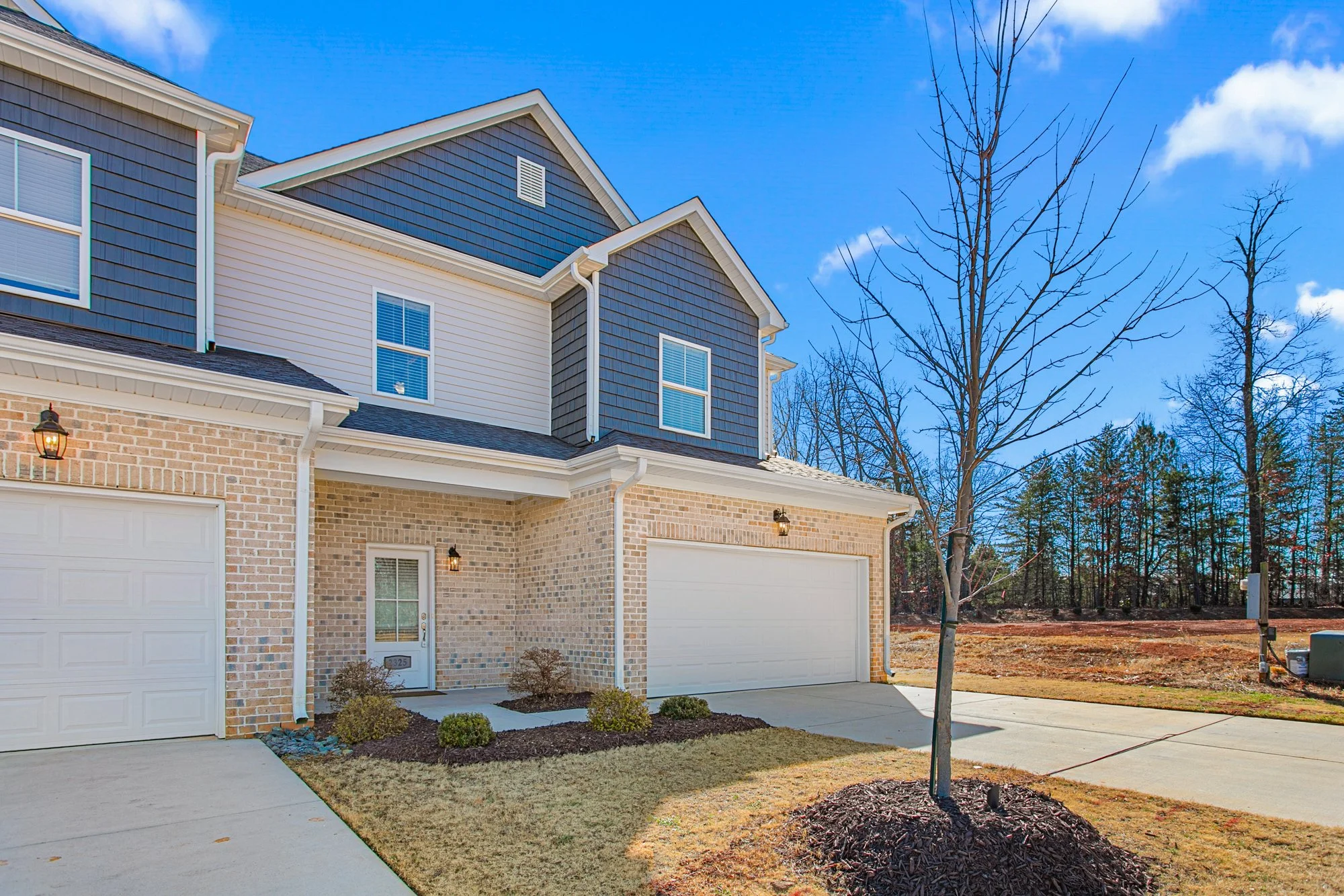 Front view of a new, two-story house with brick and siding exterior, small front yard, leafless trees, blue sky with some clouds, and a driveway.