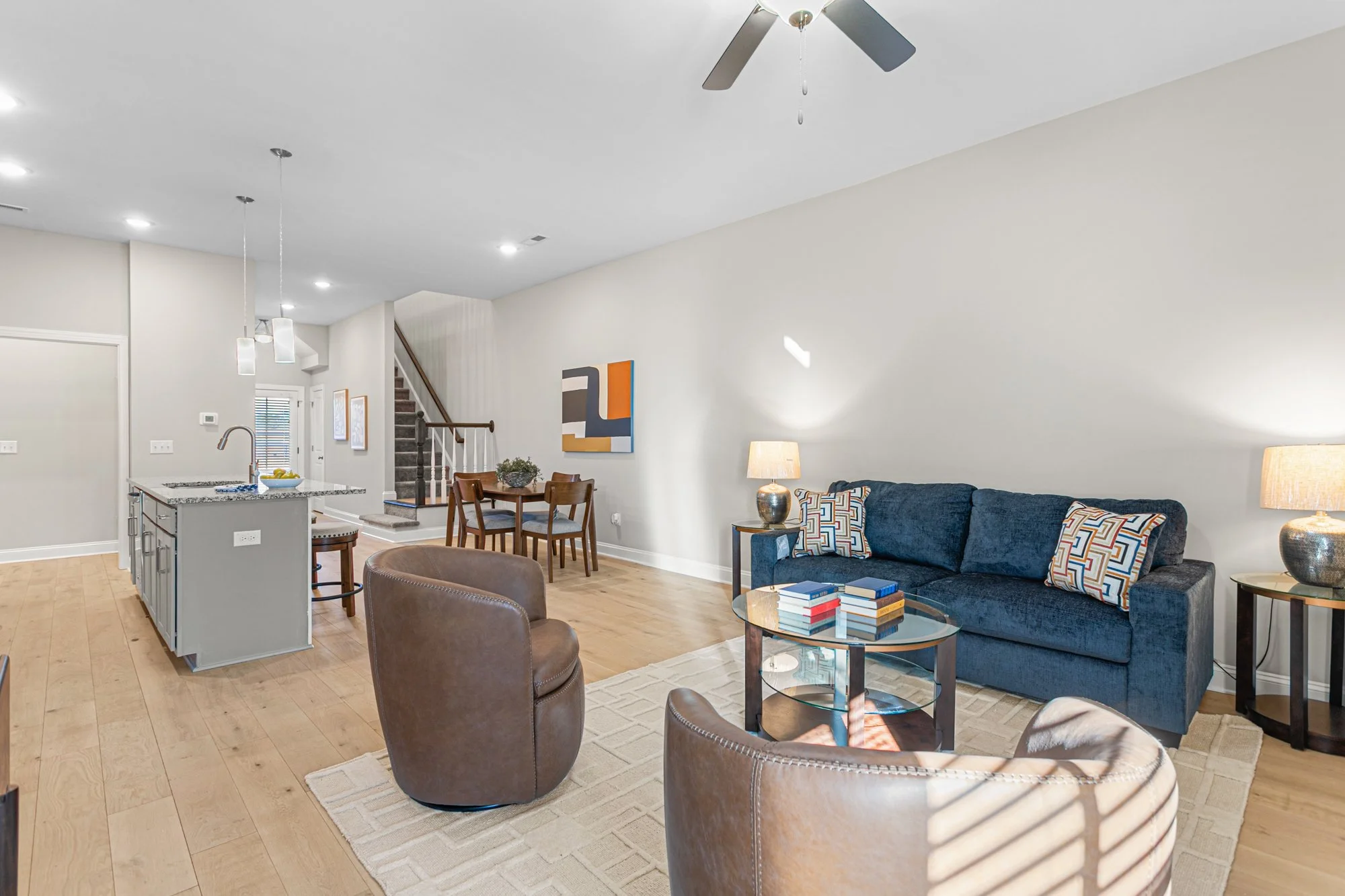 Open-concept living room with blue sofa, brown leather chairs, glass coffee table, and a dining area with a wooden table and chairs, with artwork on white walls, hardwood floors, and ceiling fans.