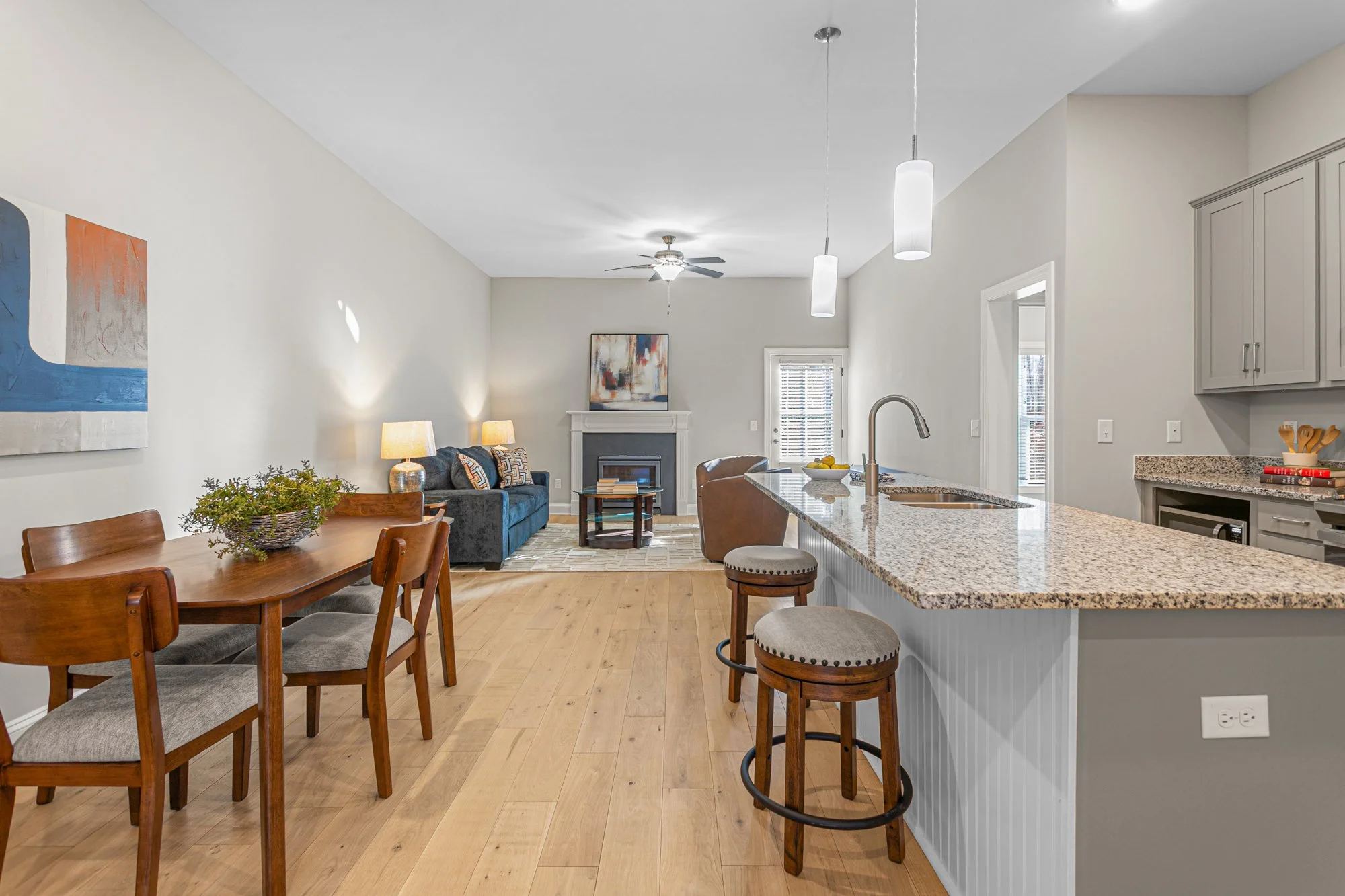 Open-concept living room and kitchen with wooden dining table, gray kitchen cabinets, granite countertops, and bar stools, with a fireplace and ceiling fans.