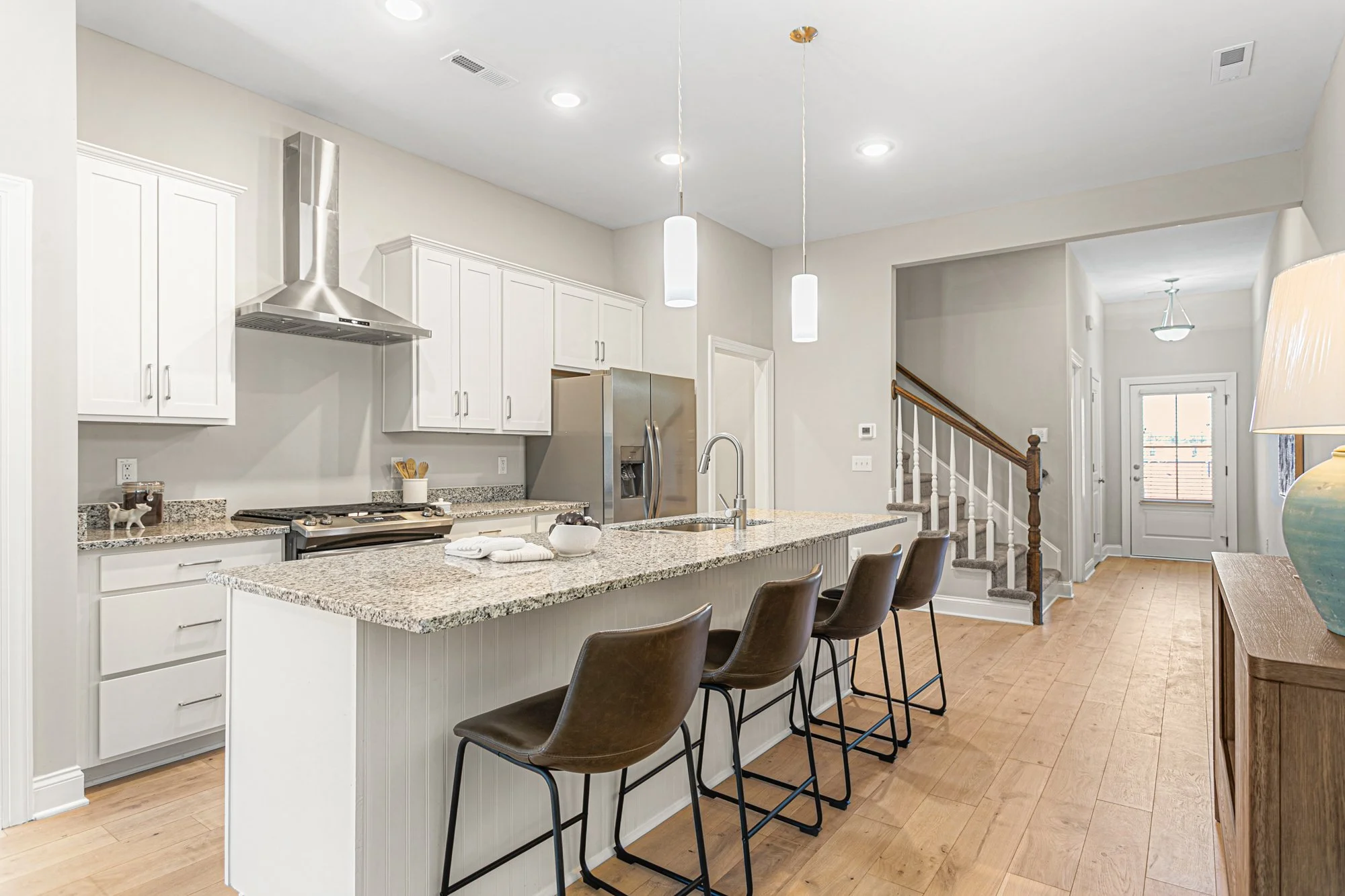 Modern kitchen with white cabinets, granite countertops, stainless steel appliances, island with three brown chairs, hardwood flooring, and a staircase leading to the upper level.