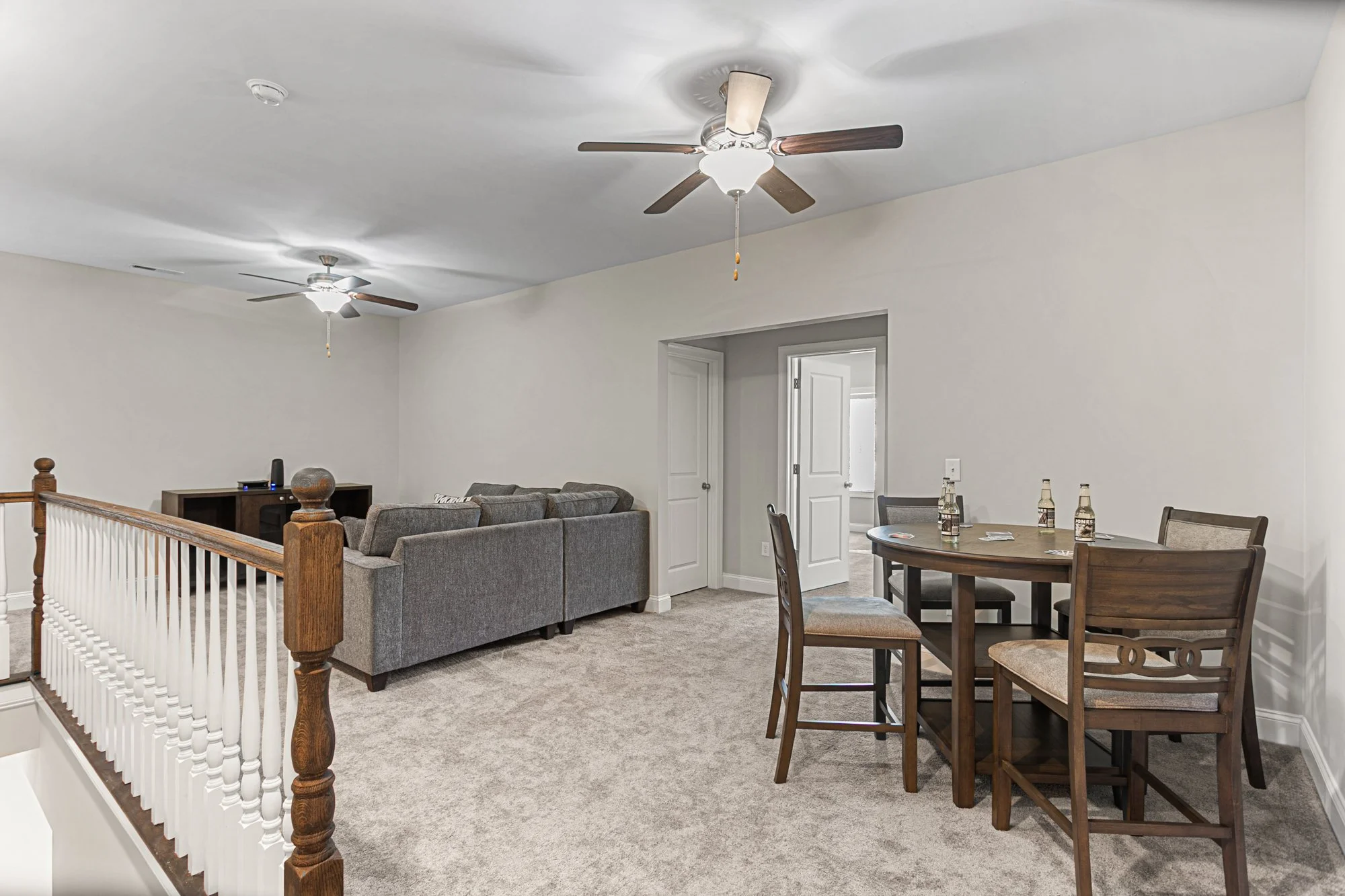 Living room with gray sofa, round wooden dining table, four chairs, ceiling fans, and beige carpet.