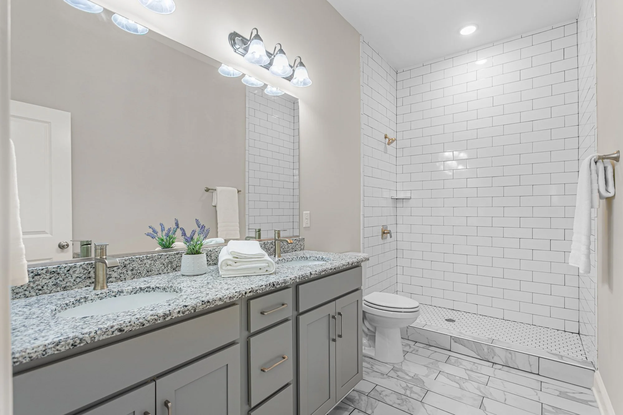 A modern bathroom with a granite countertop double vanity, two sinks, decorative towels, lavender plants, a large mirror, a white toilet, and a walk-in shower with white subway tiles and marble floor tiles.