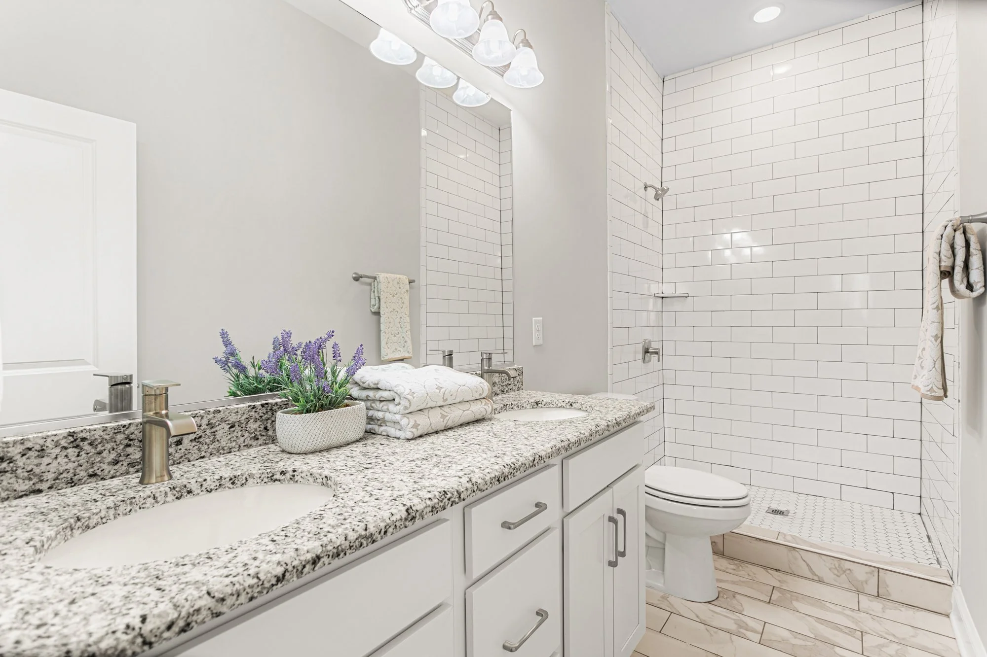 Bathroom with white cabinets, granite countertop, two sinks, a large mirror, a potted plant, towels, and a walk-in shower with white subway tiles.
