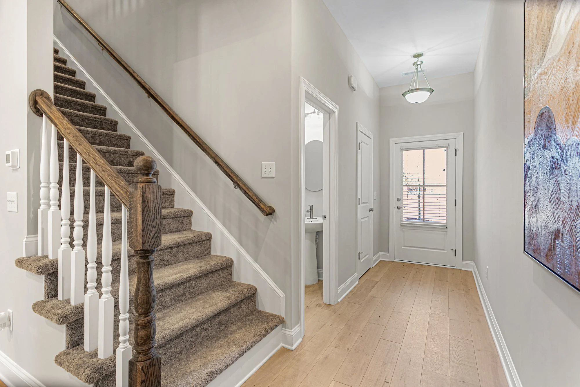 A hallway with a staircase, a door with a window, a small sink, and a large abstract painting on the wall.