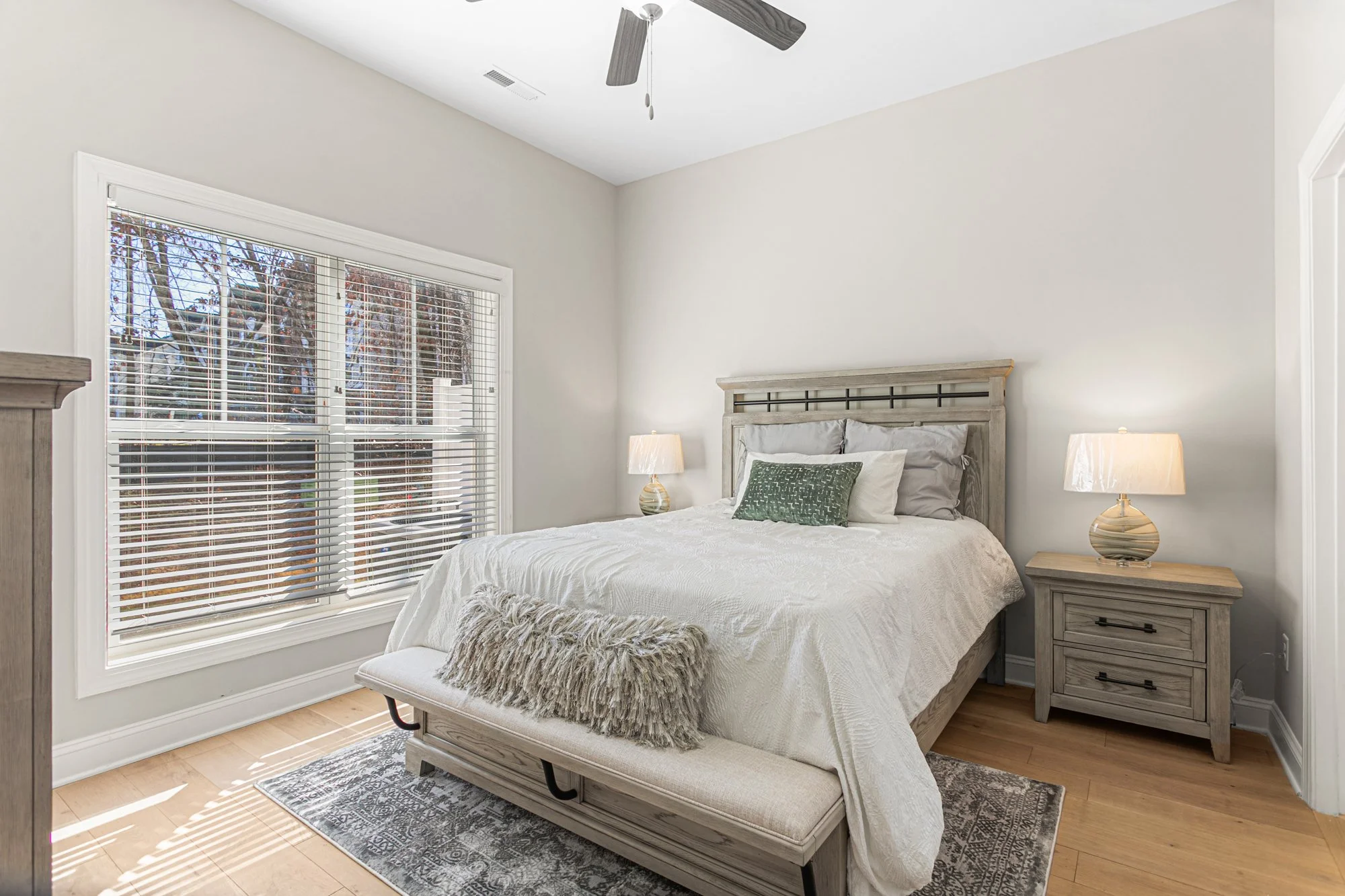 A minimalist bedroom with a large window, a wooden bed with white bedding, and matching nightstands with lamps. There is a bench at the foot of the bed with a furry pillow on it.