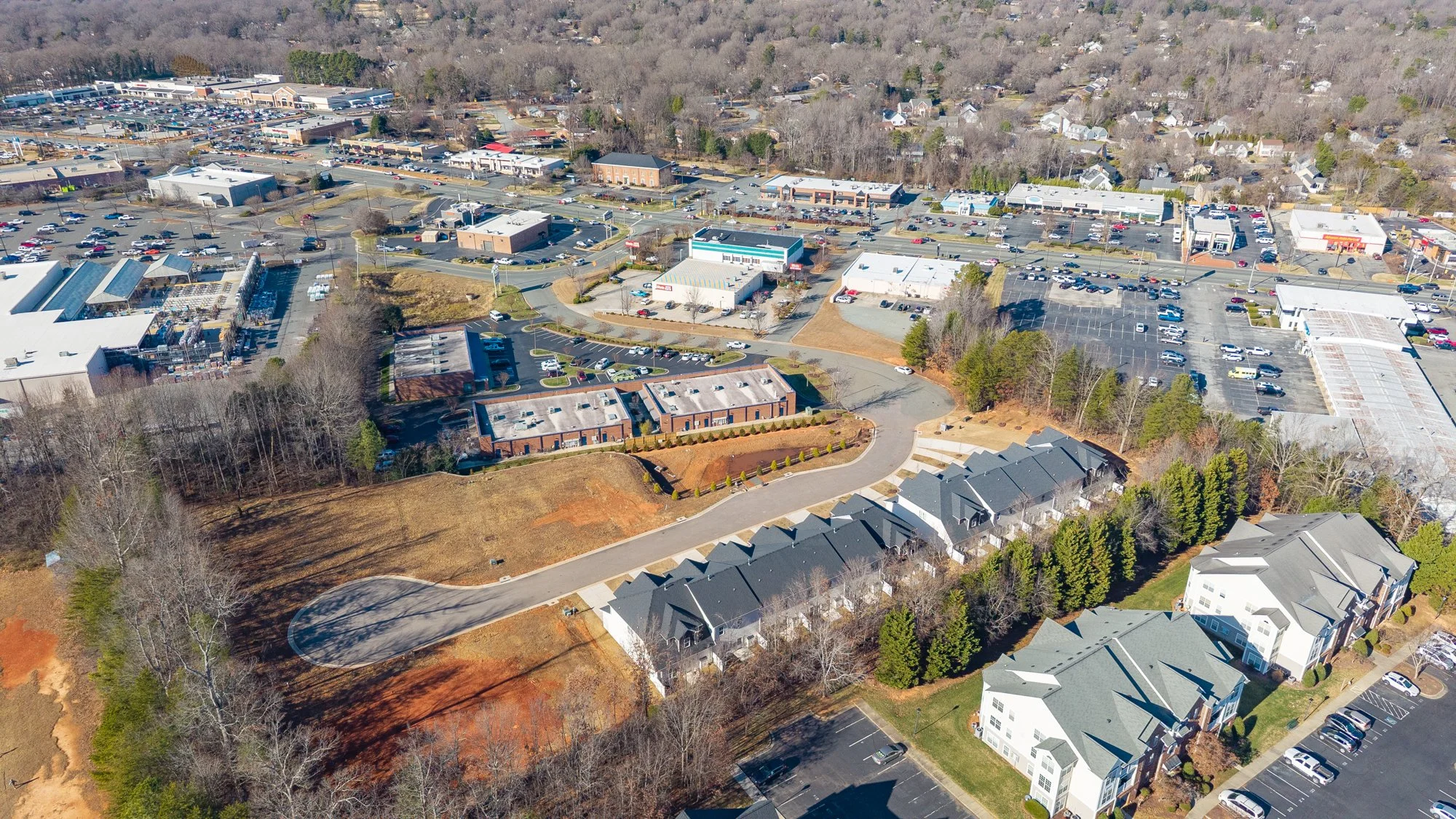 Aerial view of a shopping center, parking lots, commercial buildings, residential townhomes, and wooded areas.