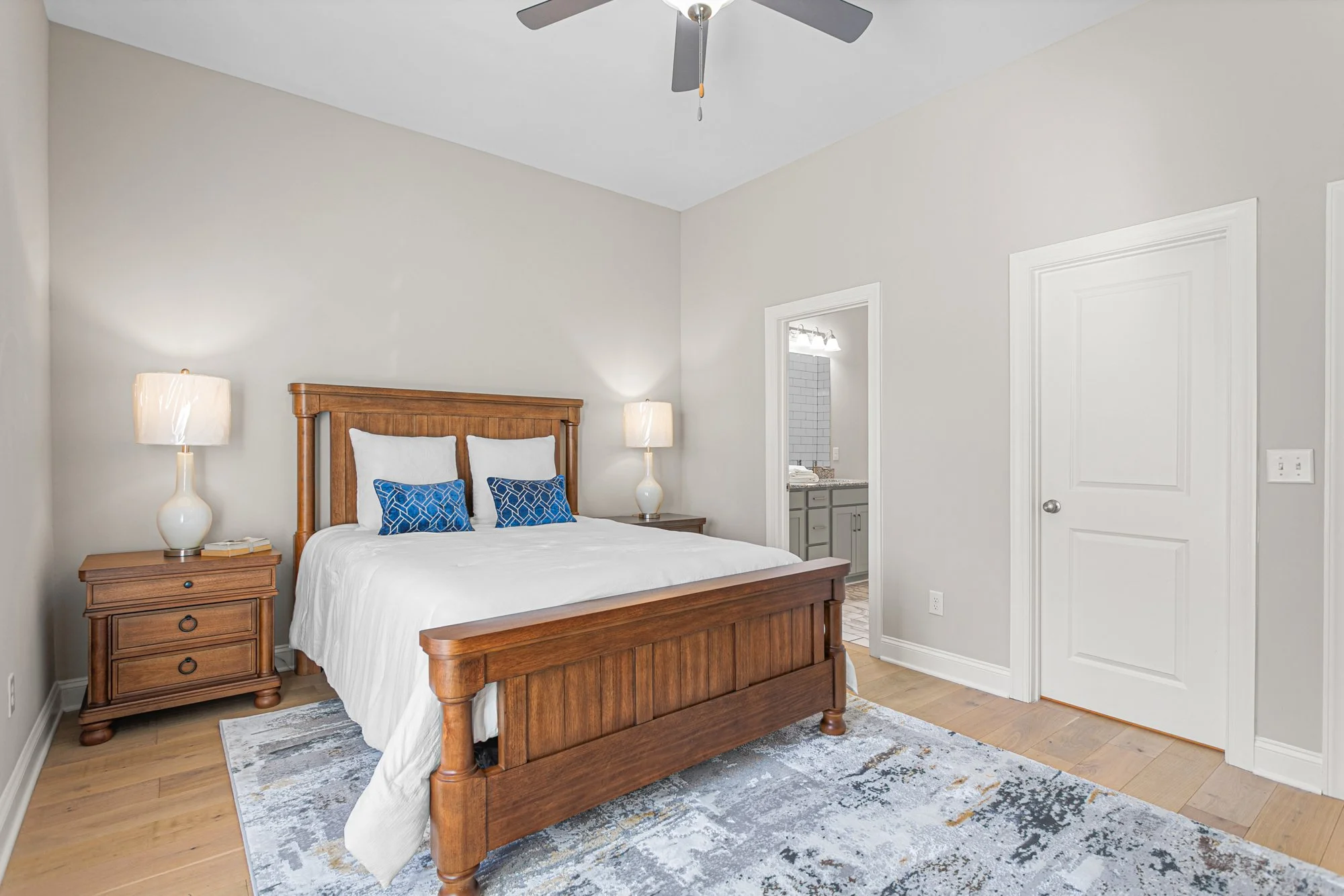 A bedroom with a wooden bed frame, white bedding, and four pillows, two white and two blue with a geometric pattern. Two matching wooden nightstands with lamps sit on either side of the bed. An area rug lies under the bed on light-colored hardwood fl