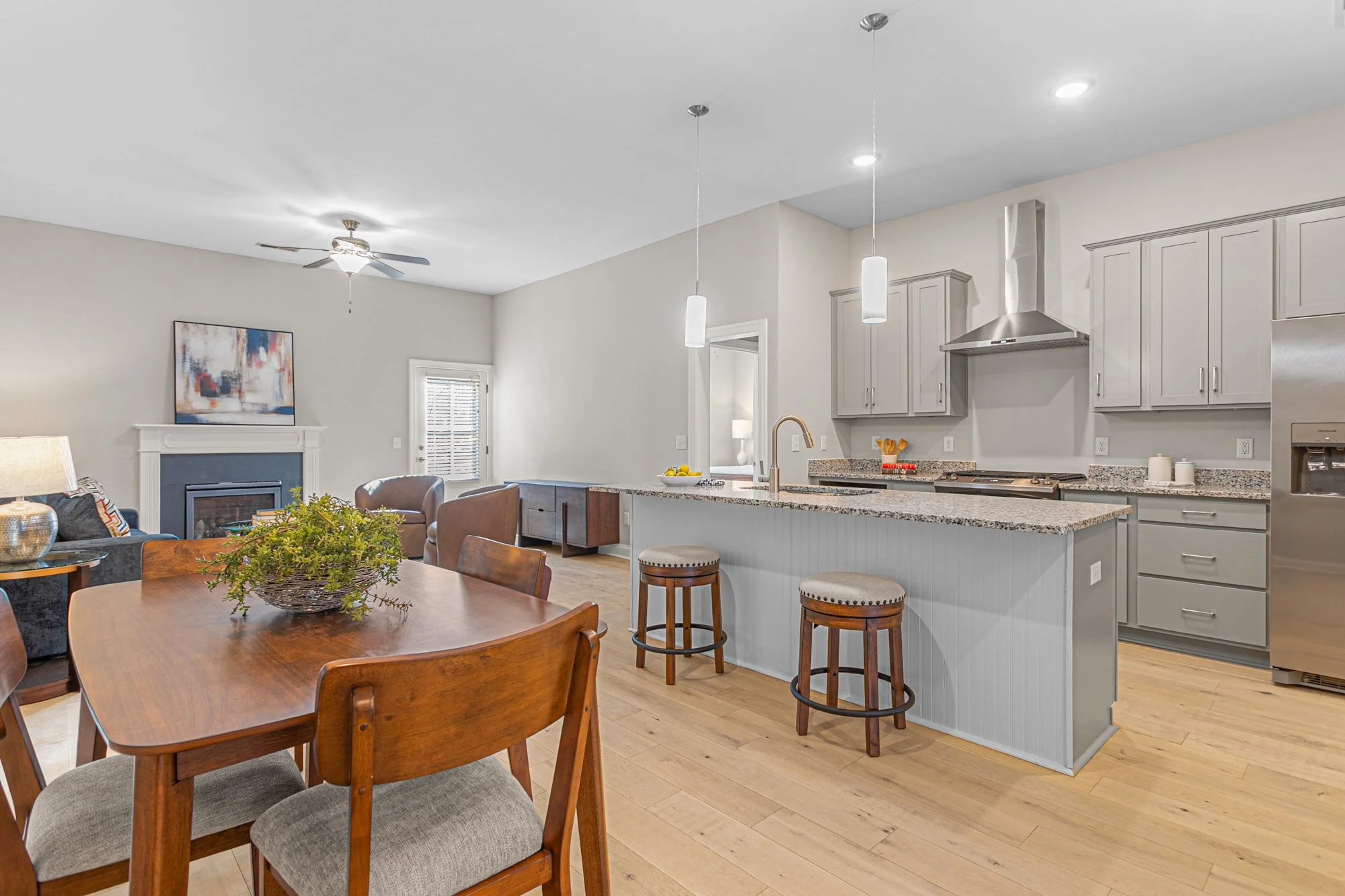 Open-concept kitchen and dining area with light gray cabinets, granite countertops, stainless steel appliances, and a wooden dining table with chairs. The space includes a living room with a fireplace, a ceiling fan, and a small window.