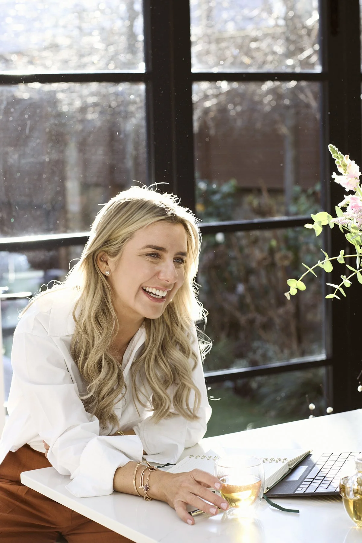 Lainey Corcoran wearing a white blouse, sitting at a table with a glass of water, an open notebook, a laptop, and a small vase with flowers, in front of a large window with sunlight streaming in.