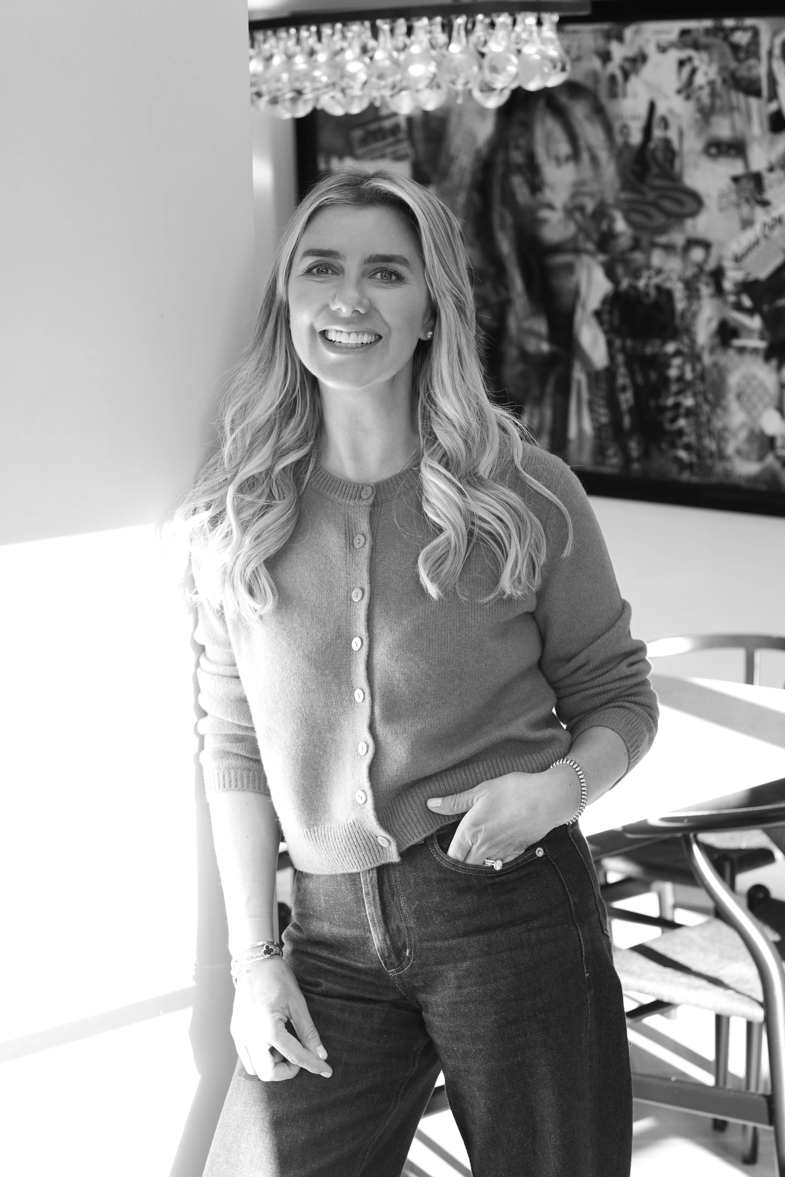 Lainey smiling stands indoors near a table, wearing a cardigan and jeans, with artwork in the background.