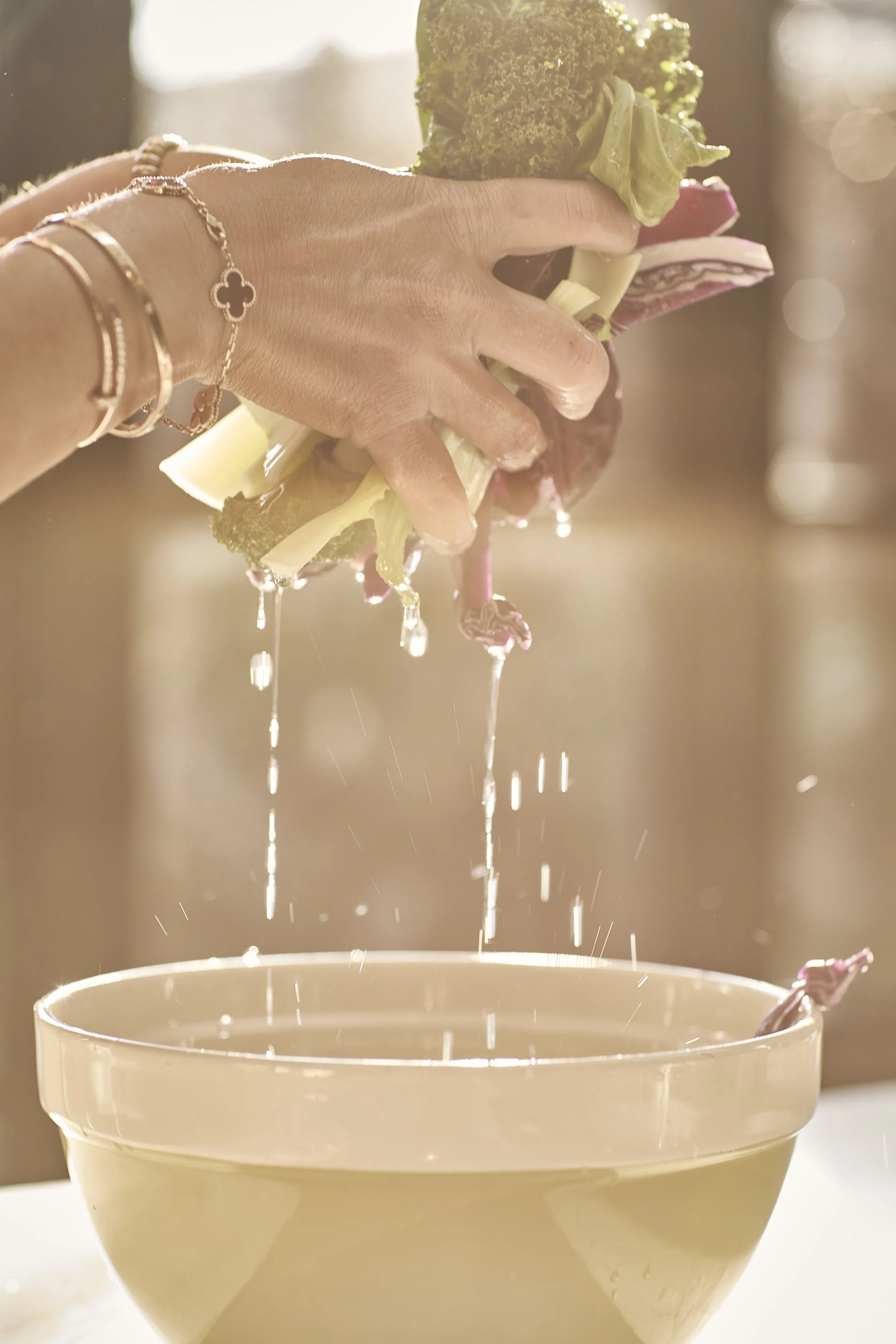 A hand holding a bunch of fresh vegetables, including broccoli, leeks, and radicchio, above a large mixing bowl, with water dripping from the vegetables, in natural sunlight.