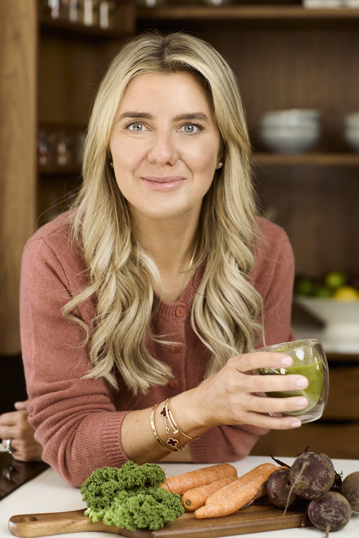 Smiling at the camera, sitting at a kitchen counter with fresh vegetables including carrots, beets, and broccoli in front of her. She is holding a glass bowl with a green smoothie inside and wearing a pinkish-red sweater with jewelry on her wrist.