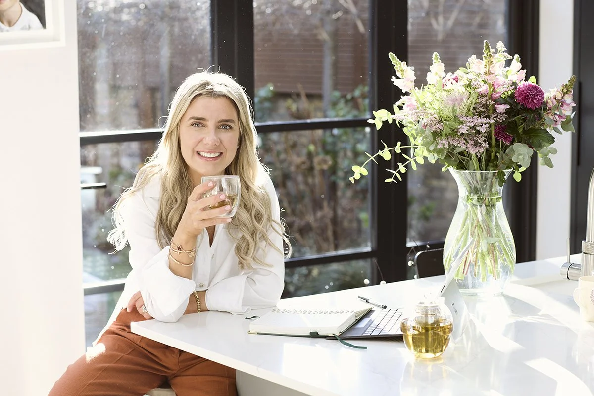 Lainey smiling and holding a glass of water in a bright kitchen with large windows, a bouquet of pink and purple flowers in a glass vase, an open notebook, and a laptop on the white countertop.