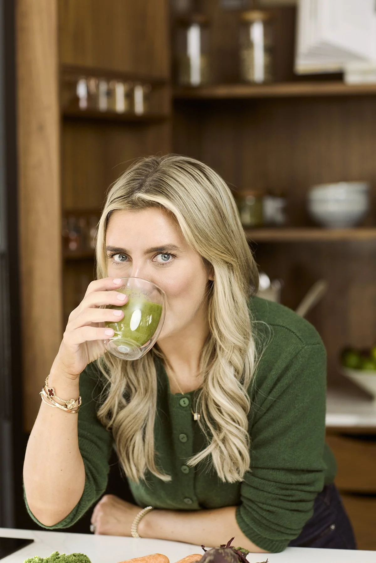 Lainey wearing a green sweater, drinking a green juice from a clear glass in a kitchen.