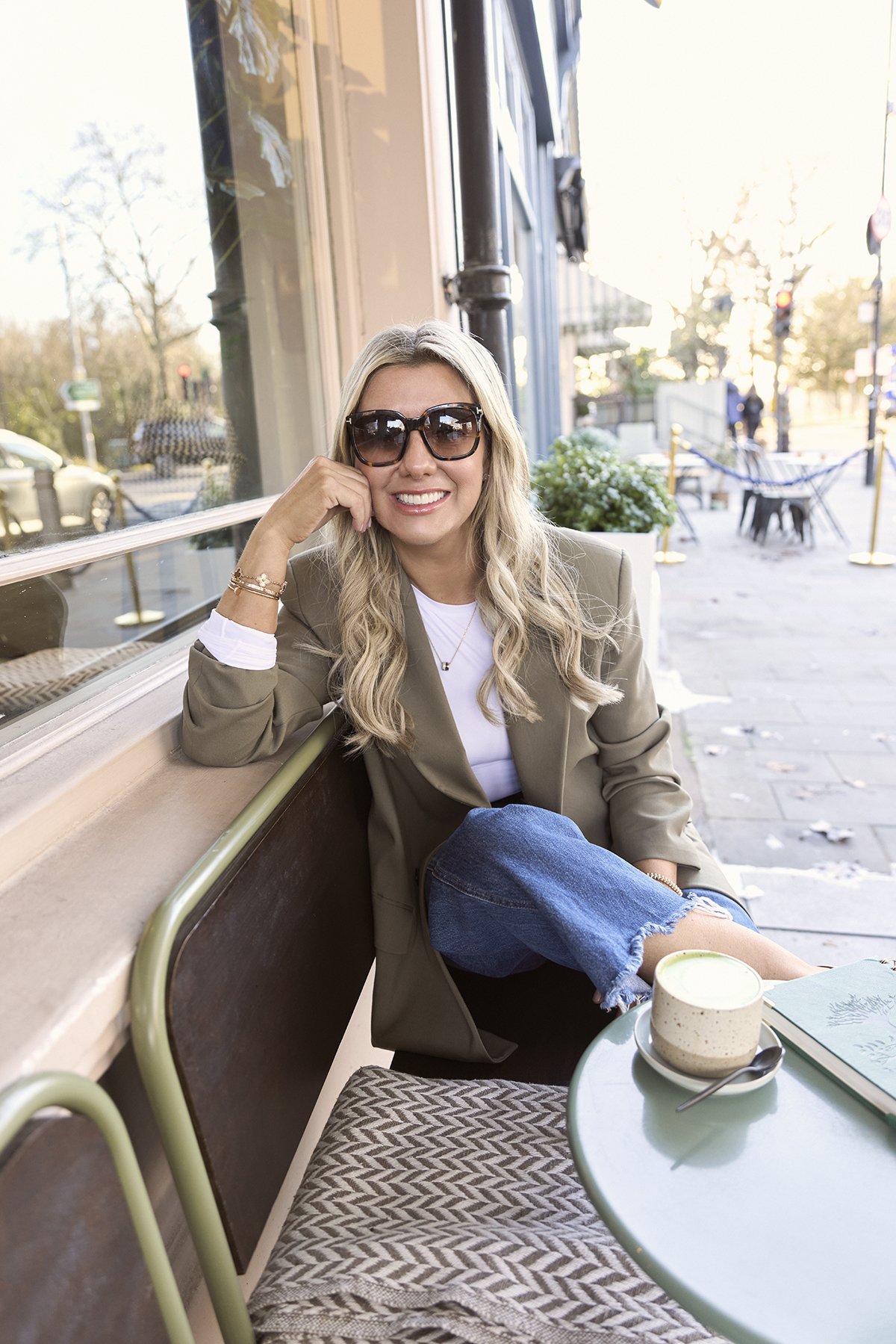 Lainey sitting outside at a cafe, wearing a beige blazer, white shirt, and ripped jeans, with a cup of tea or coffee on the table beside her.