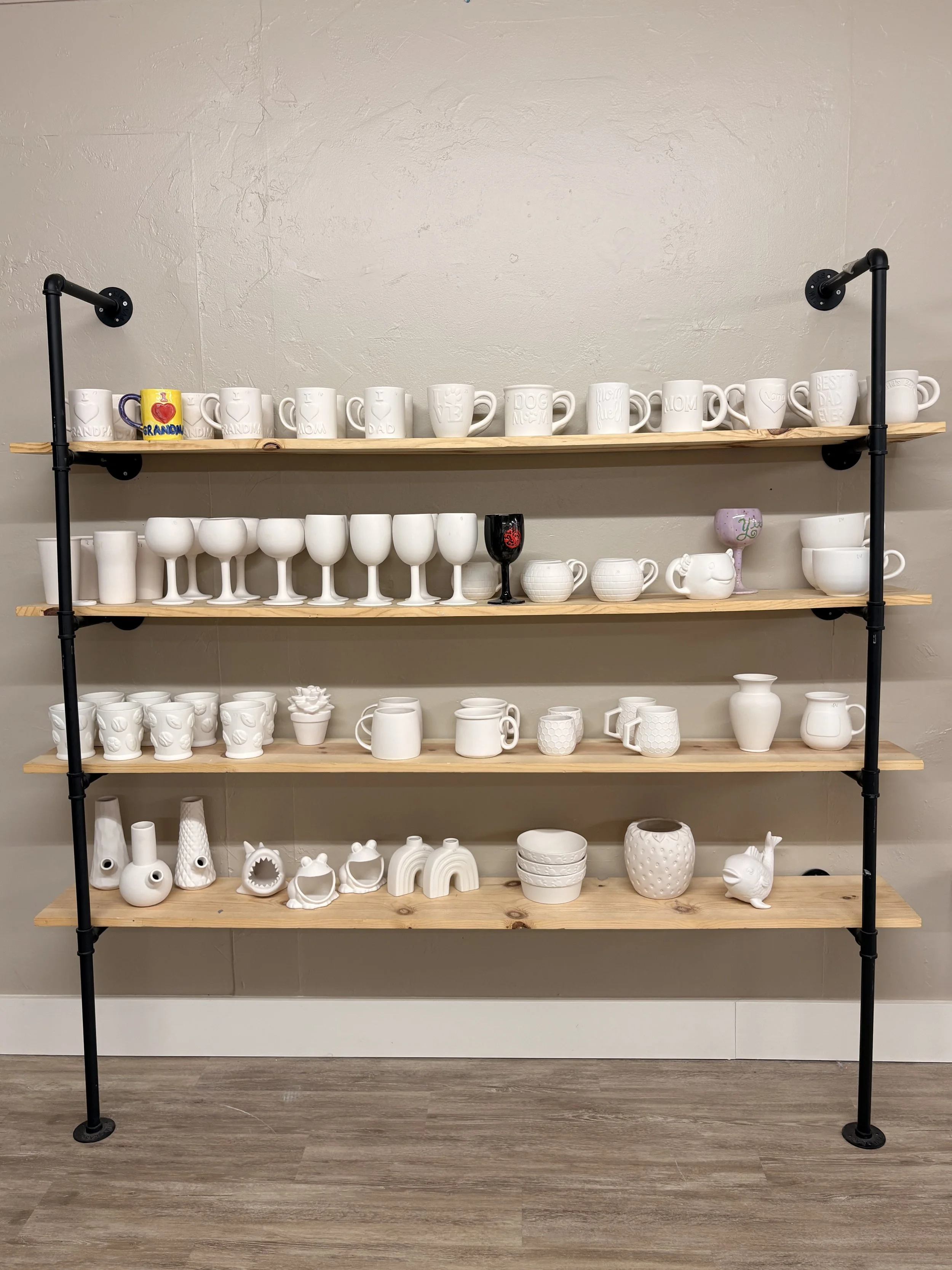 Wooden shelf with various white ceramic mugs, cups, and decorative items, some with handles, some without, on a black metal frame against a beige wall.