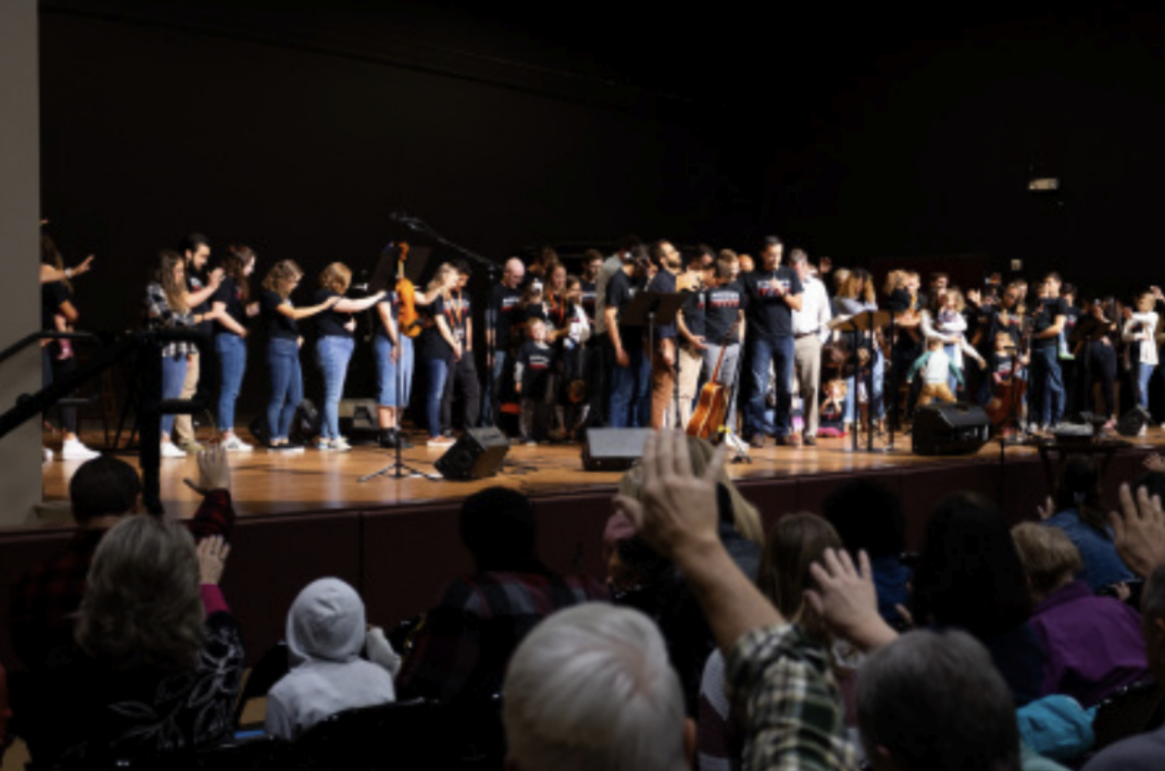 A large group of people on stage, some holding instruments, taking a bow after a performance, with an audience clapping and raising their hands in front of the stage.