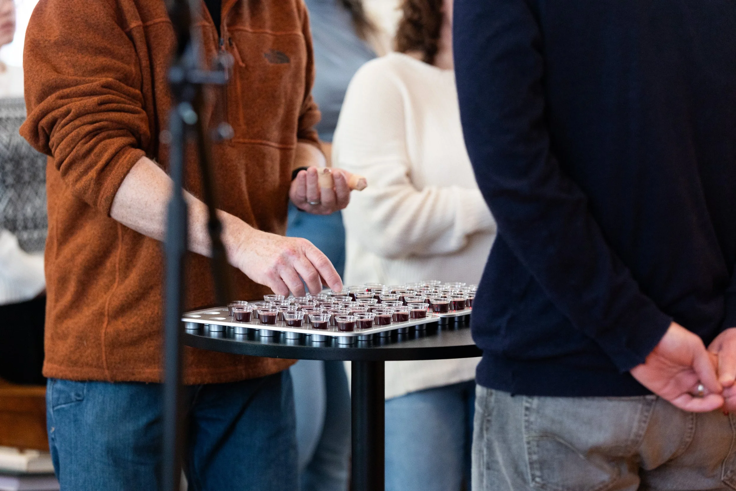 Close-up of a person in a brown fleece jacket holding a spoon, standing near a tray of small glasses filled with dark red liquid, with other people blurred in the background.