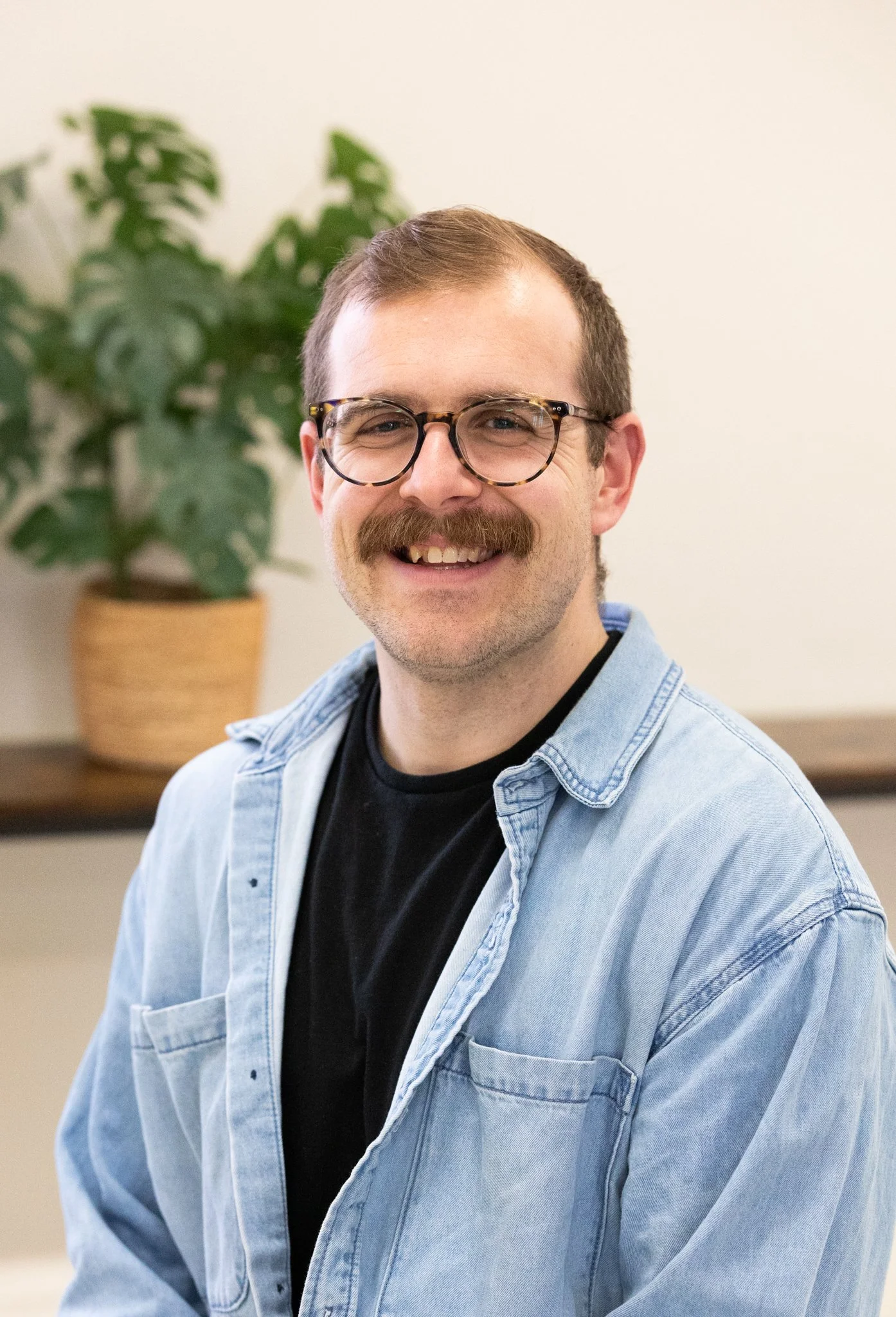 Smiling man with glasses and mustache wearing a denim shirt over a black t-shirt, sitting in front of a beige wall with a potted plant in the background.