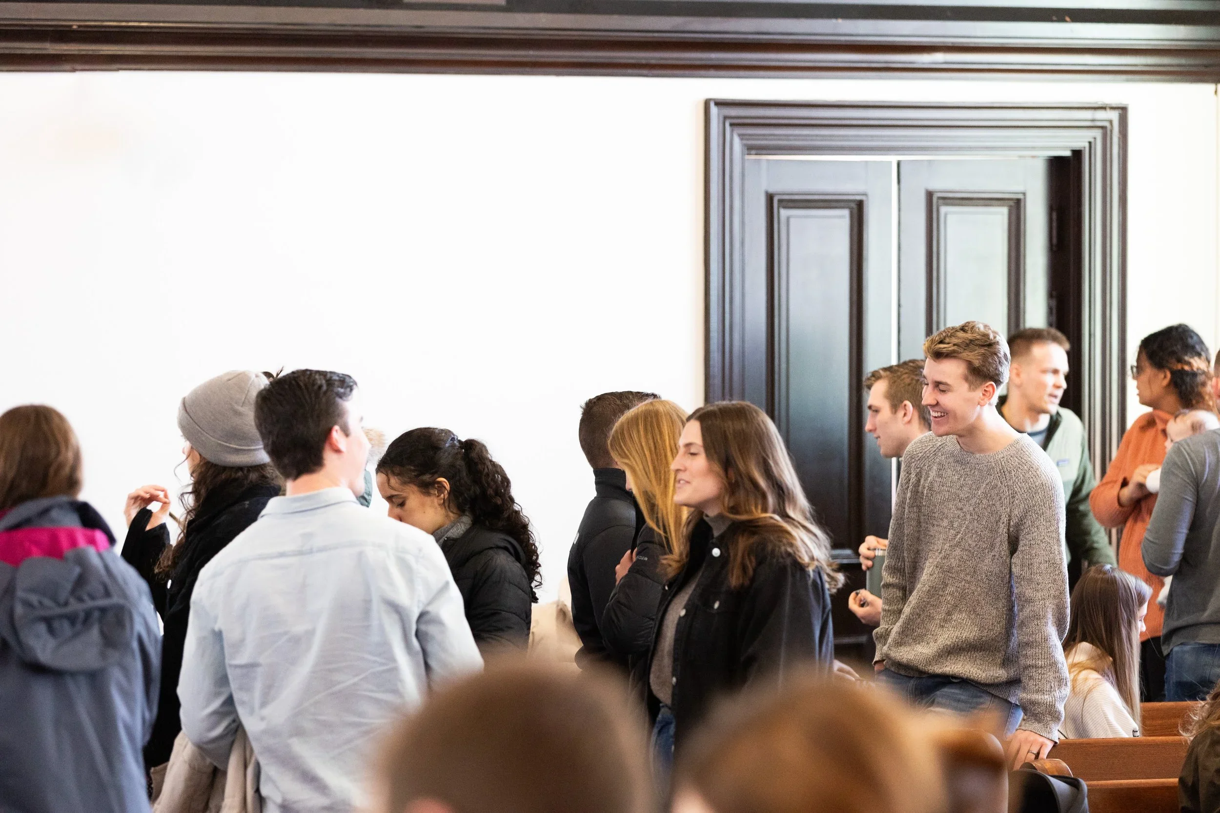 People standing in line inside a building, some engaging in conversation, with a large dark wooden door in the background.