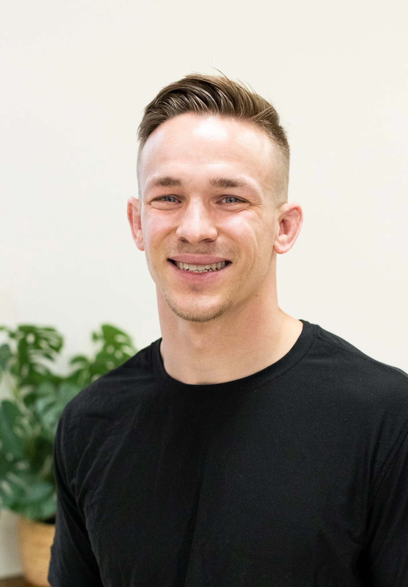 A smiling young man with short, styled hair wearing a black t-shirt standing indoors with a light-colored wall and a green leafy plant in the background.