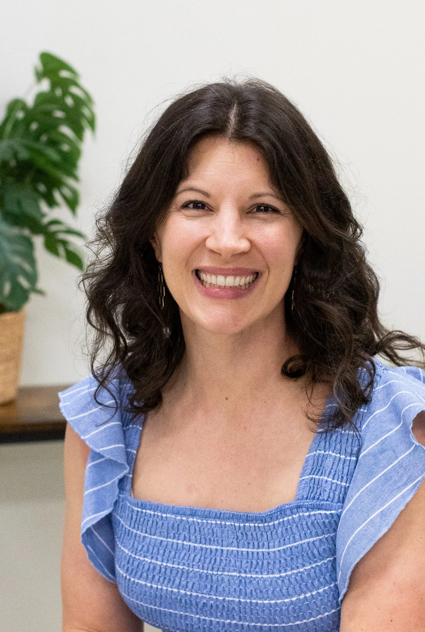 A woman with dark wavy hair smiling, wearing a blue ruffled sleeveless top, in front of a plain white background with a potted plant on a table to her side.