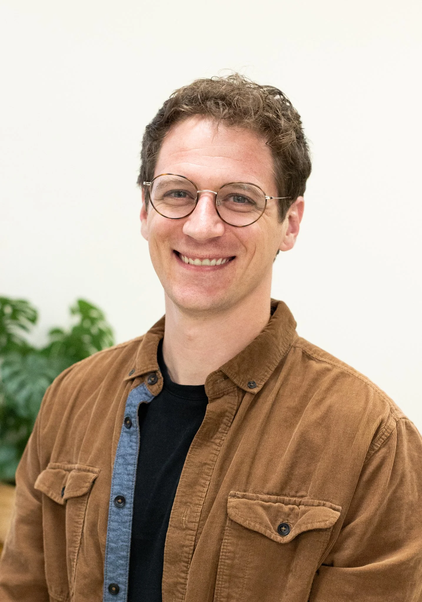 A smiling man with curly hair, glasses, wearing a brown jacket over a black shirt, standing against a plain white background with a green plant in the background.