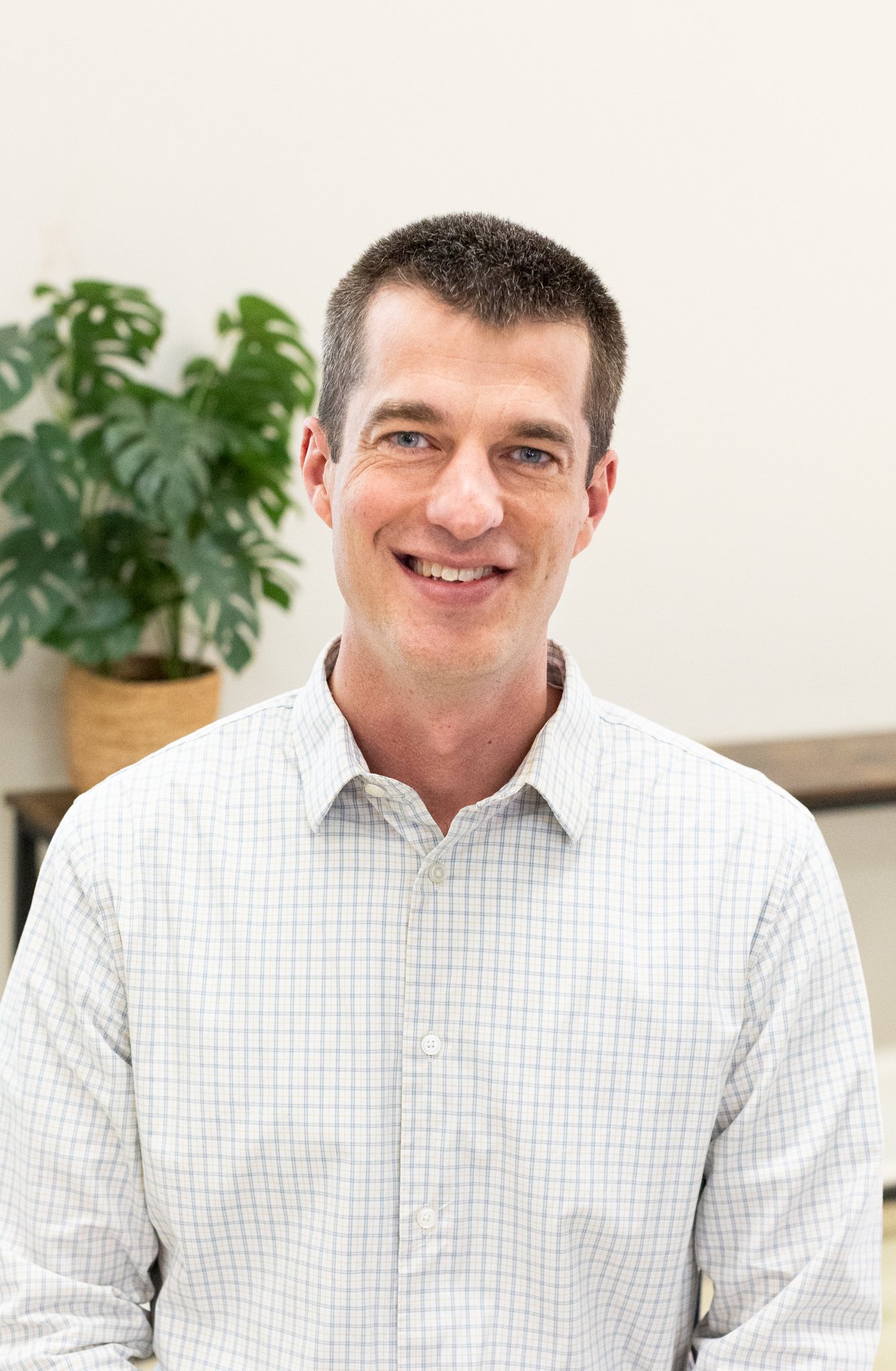 A smiling man with short dark hair wearing a light-colored, checked button-up shirt, standing in front of a potted plant and neutral-colored wall.