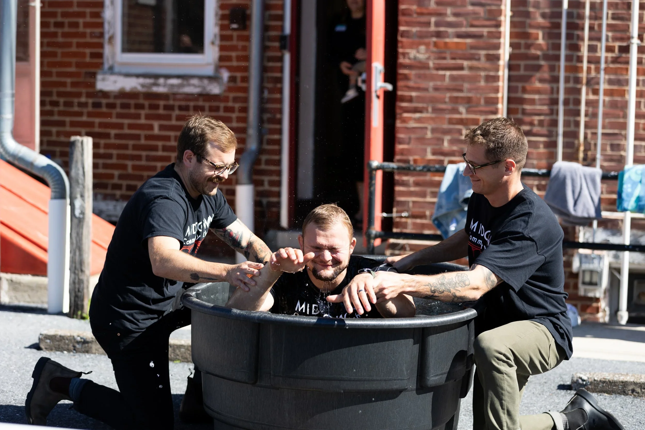Three men, two kneeling and one sitting in a black plastic tub outside, are participating in a baptism. They are smiling and holding the man in the tub as water splashes around. In the background, a brick building with a window and laundry hanging outside is visible.