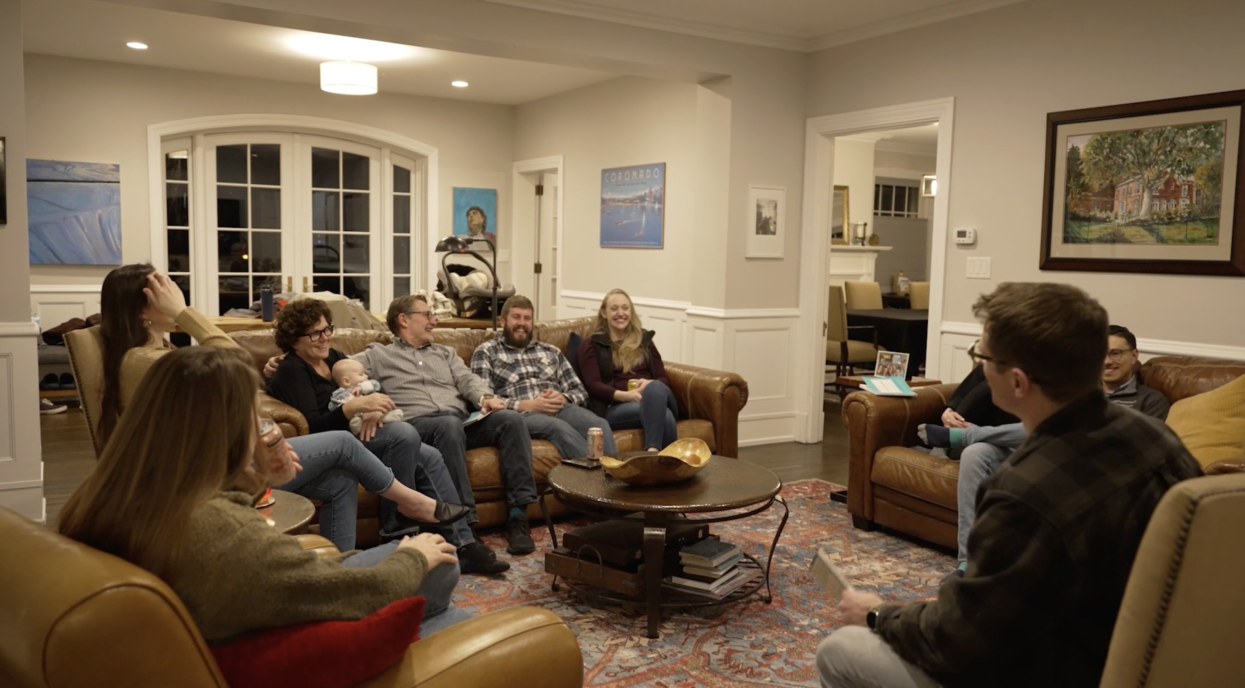 A group of people sitting on couches and chairs in a living room, engaging in a social gathering, with some smiling and laughing, while a baby is held by a woman.
