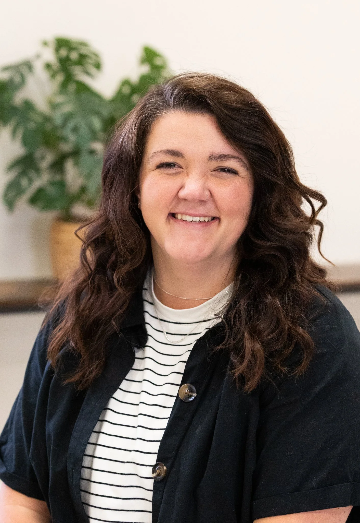 A woman with long, wavy brown hair smiling at the camera, wearing a black shirt over a white and black striped top, with a plant in the background.